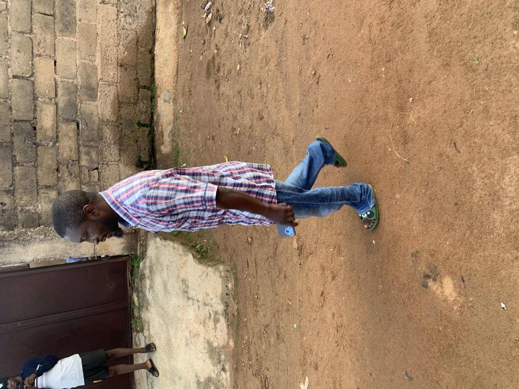 In Gura Top, Nigeria, a man is walking sideways on a dirt path next to a weathered brick wall. He is wearing a red and blue plaid shirt, blue jeans, and green slides with cartoonish eye details. He is holding a dark-colored object, possibly a phone, in his right hand. To the left of the frame, another person stands near a large brown metal door, wearing a white t-shirt, black shorts, and black sandals. The ground is covered in dirt and small stones, with patches of dry grass and scattered debris, including a few pieces of paper and a bent piece of straw forming a rough octagon shape. The lighting suggests it is daytime.