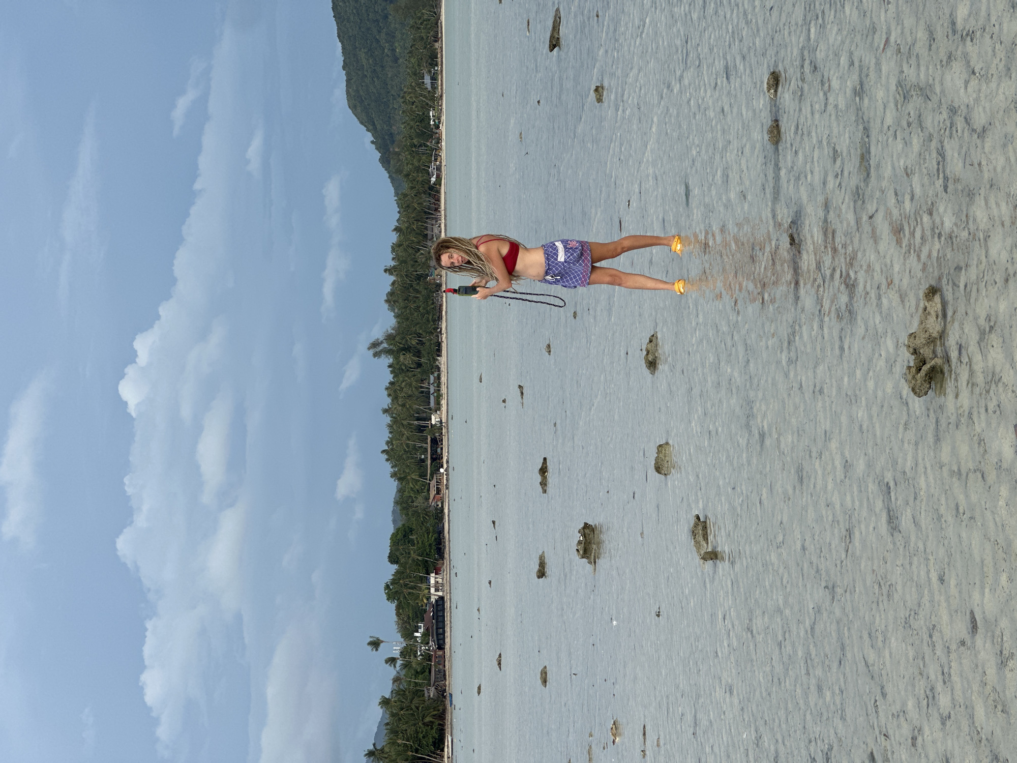 A woman with dreadlocks is standing in shallow water on a beach in ตำบลเกาะพะงัน, Thailand, holding a black object. She is wearing a red bikini top and blue and white checkered shorts with yellow sandals. The water is clear and calm, revealing a sandy bottom with scattered rocks. In the background, there is a coastline with trees and buildings. The sky is blue with some clouds.