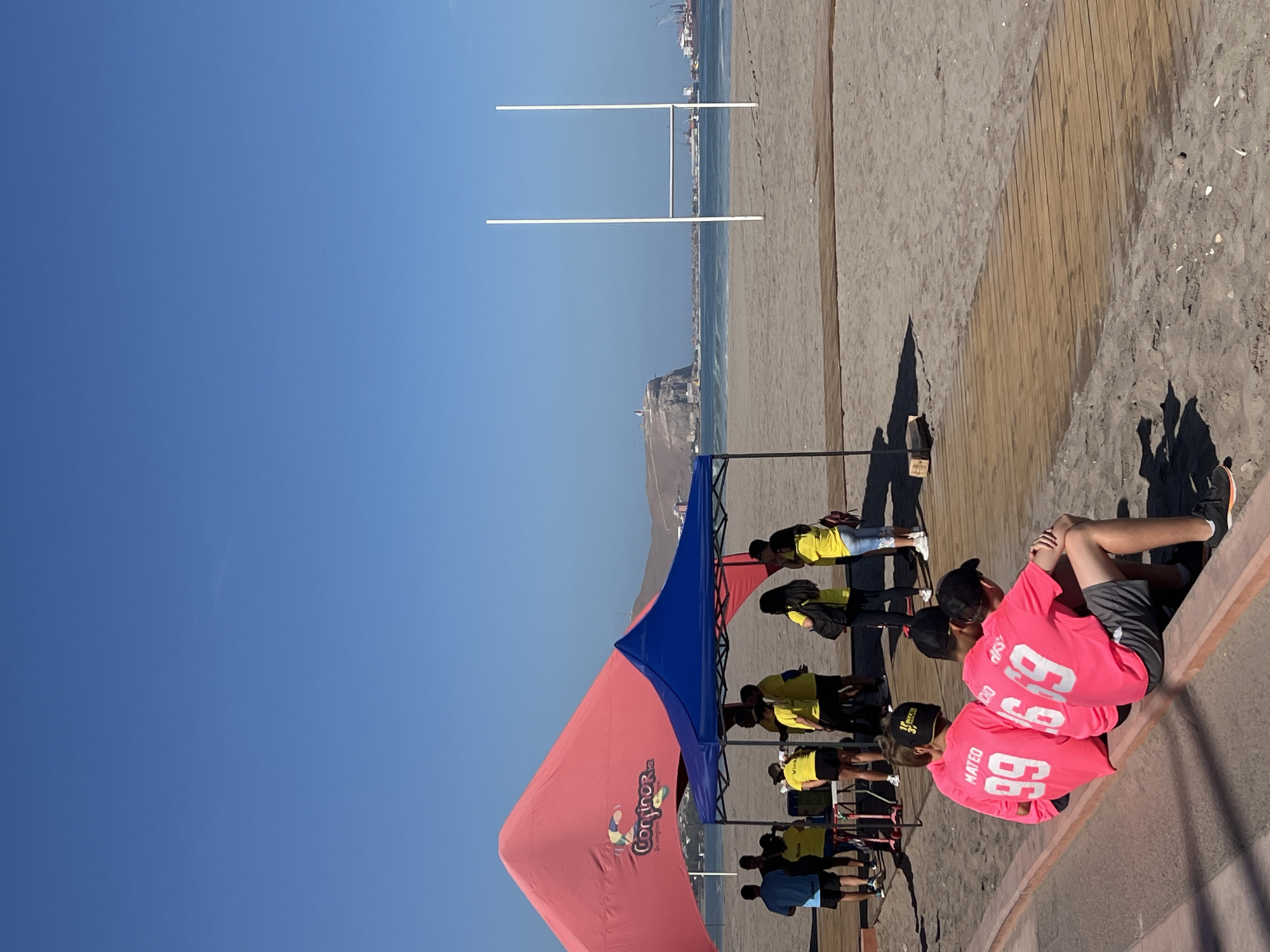 This media file captures an outdoor event taking place on a sandy beach adjacent to a wooden boardwalk, under a clear, vibrant blue sky. The setting is likely Arica, Chile, given the coastal features and port activity.
In the immediate foreground, two young individuals are seated with their backs to the camera, wearing bright pink long-sleeved shirts. Visible text on their shirts includes "MATEO 99" and another "99" with a partially obscured name, possibly "CHO".
To the left, a colorful event setup is present, featuring two pop-up canopies. One canopy is distinctively red/pink with a blue top section, displaying a logo that includes a worm-like character and the text "Congrere" or "CongriOra". The other canopy is entirely blue. Numerous people are gathered around and under these canopies, many wearing yellow shirts, suggesting they are participants or staff of an organized activity.
In the mid-ground, on the sandy beach closer to the water, two white goalposts, resembling those used in rugby or American football, are prominently set up. Beyond the beach, the ocean extends to the horizon, where distant landforms and industrial port cranes are visible, indicative of a bustling coastal city.
The bright sunlight and distinct shadows suggest it is daytime, likely mid-morning or early afternoon, with excellent weather conditions. The overall scene depicts an active, organized beach sports event or festival.