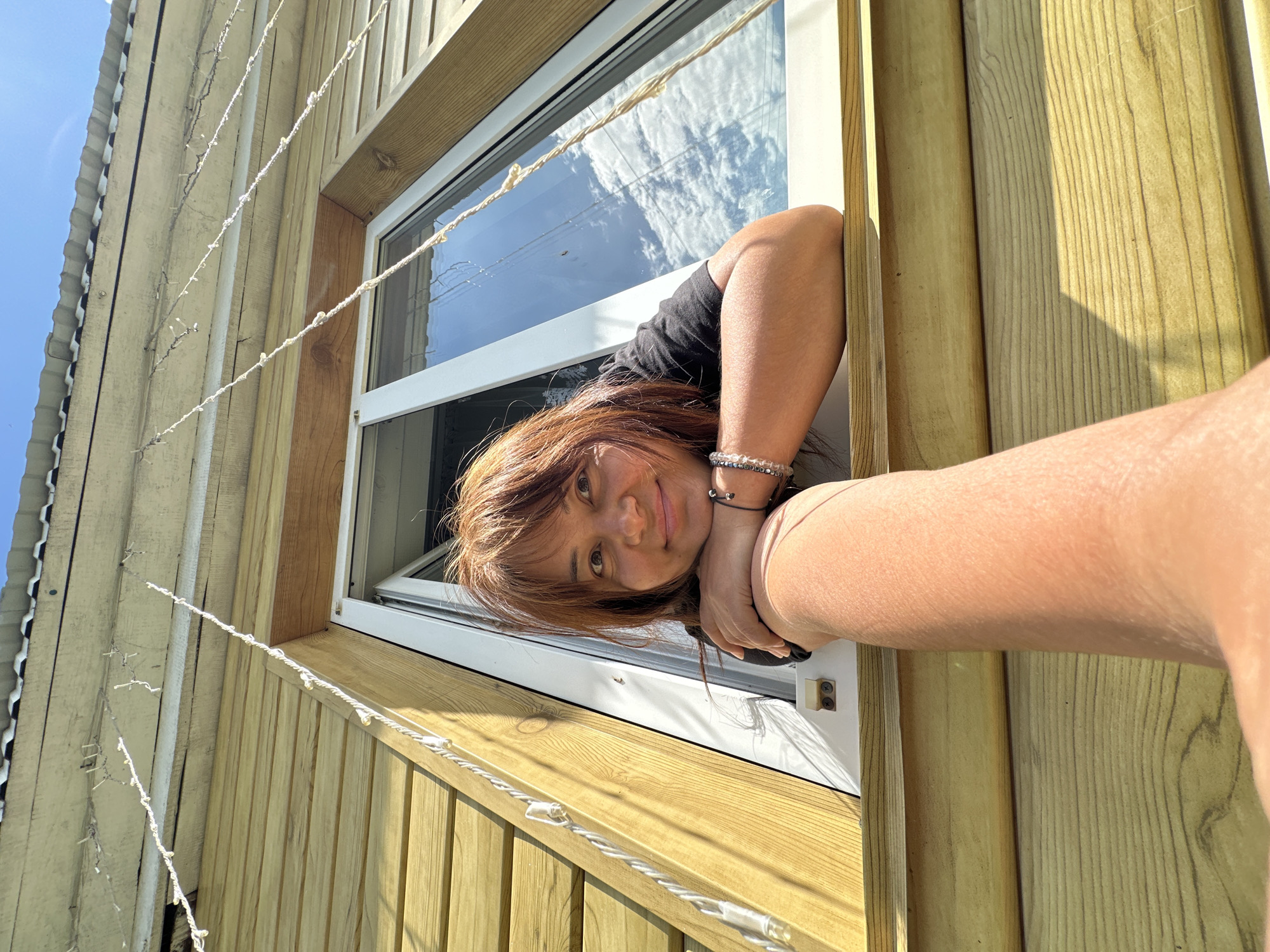 A person is leaning out of a window of a wooden house, holding their arm out towards the camera. The person has brown hair and is wearing a black shirt and a bracelet. The house is made of light-colored wood and has white window trim. There are white string lights hanging from the roof of the house. The sky is blue with some clouds. The image is taken from a low angle, looking up at the person.