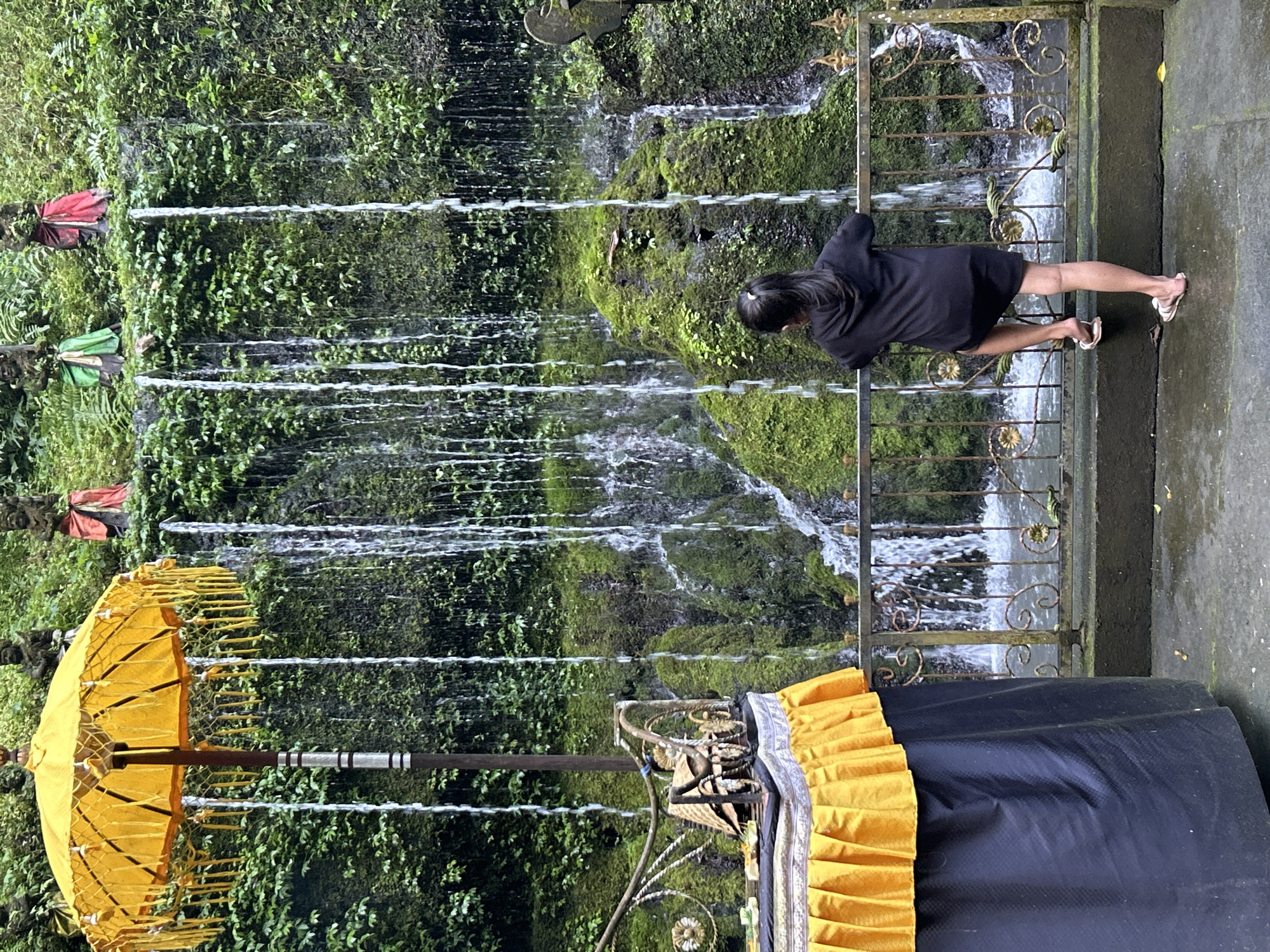A young girl in a black hoodie dress and flip-flops leans on a decorative metal fence, looking at a moss-covered rock face with multiple thin waterfalls cascading down it. In the background, three stone figures adorned with colorful cloths stand among lush green foliage atop the rock face. To the left, a large, ornate yellow umbrella with decorative tassels stands next to a table draped with a black cloth and a yellow pleated fabric skirt. The ground is wet, suggesting recent rain or spray from the waterfalls. The scene appears to be in an outdoor, natural setting, possibly a temple or sacred site, given the presence of statues and offerings. This location is identified as the Tirta Empul Temple in Bangli, Indonesia.