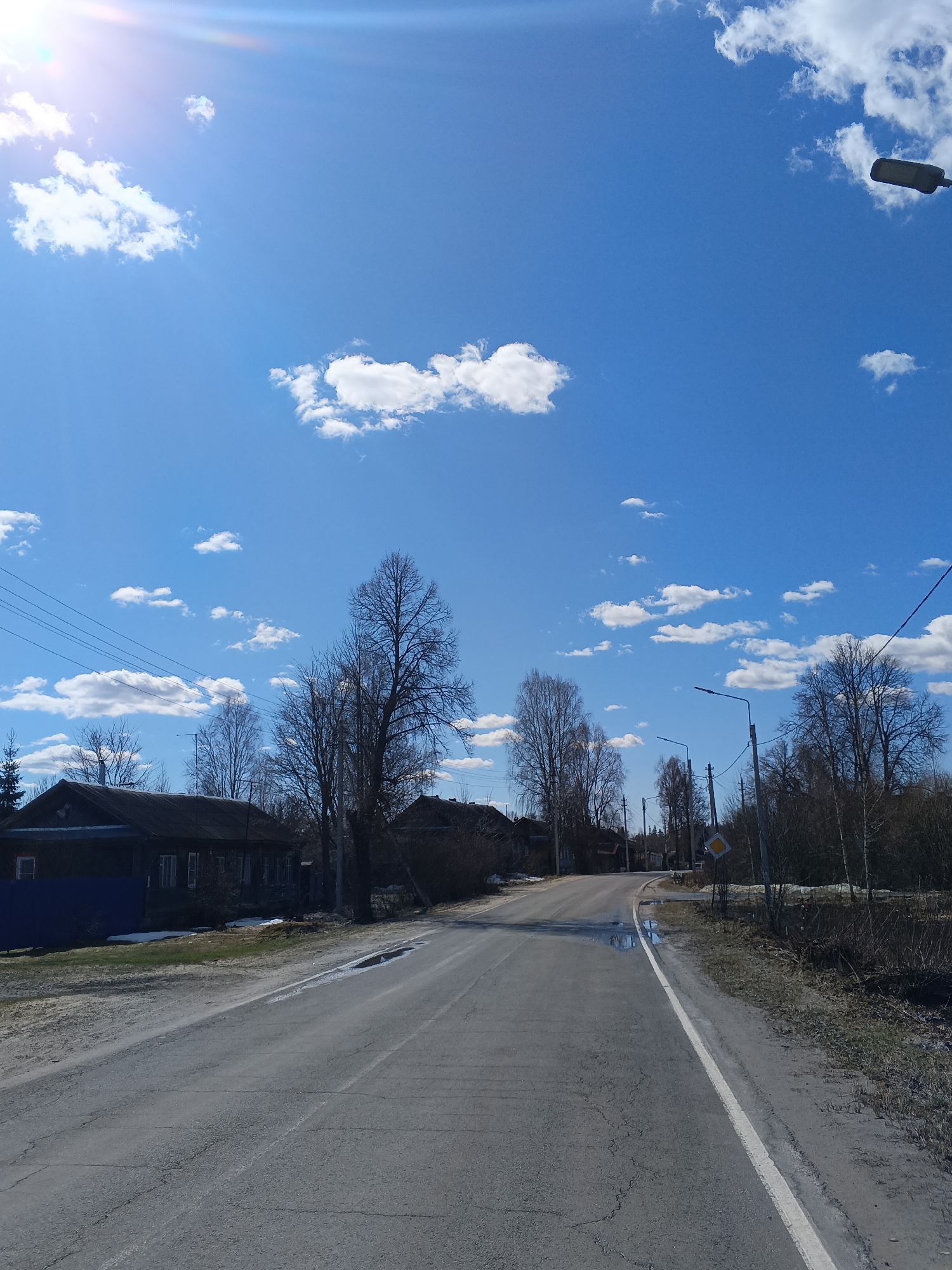 The photograph shows a street scene in Красный Маяк, Russia. The road is asphalt and appears to be moderately damaged, with cracks and patches visible. There are buildings on either side of the road, likely residential houses. The buildings are mostly made of wood and are dark in color. Trees line the road, most of which are bare, indicating it is early spring or late fall. The sky is bright blue with scattered white clouds. A street light is visible on the right side of the road. A traffic sign is also visible on the right side of the road. Patches of snow remain along the sides of the road. There are no people visible in the image. The scene seems quiet and rural.