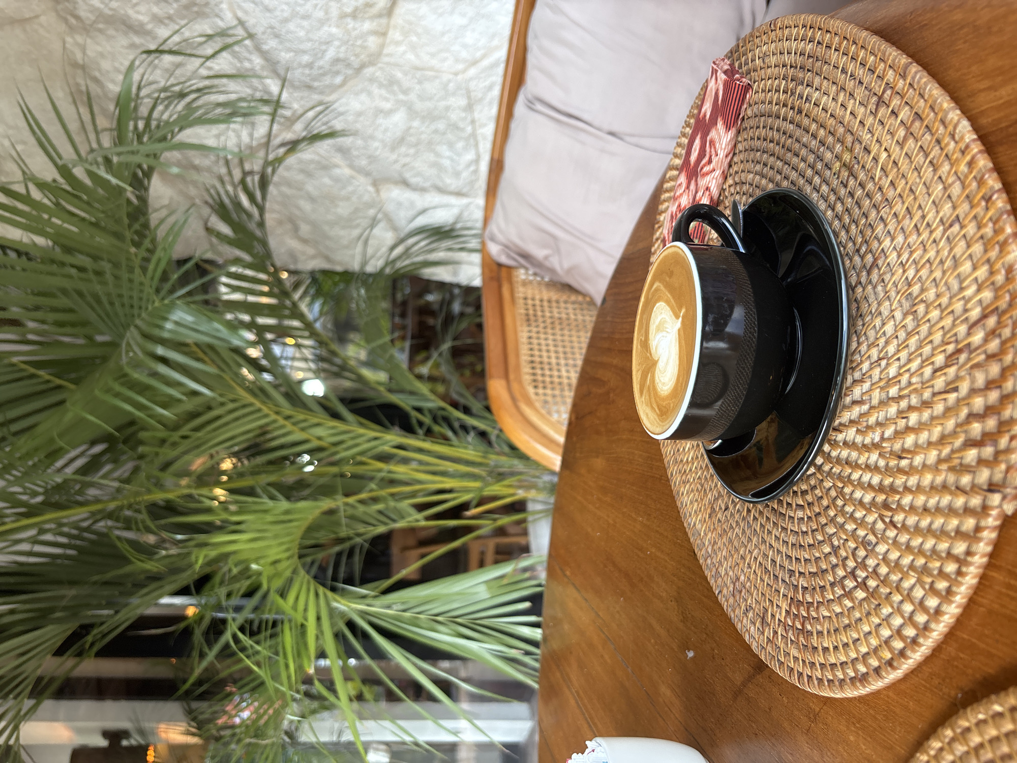 The image depicts a table setting with a cup of coffee at a cafe in Kuta Selatan, Indonesia. A black coffee cup and saucer rest on a round woven placemat on a wooden table. A folded red and white patterned cloth is visible beside the cup. To the left, a leafy green plant is present, with a woven chair and cushions in the background. The scene appears to be indoors, with a window view blurred out of focus, showing what looks to be city lights.