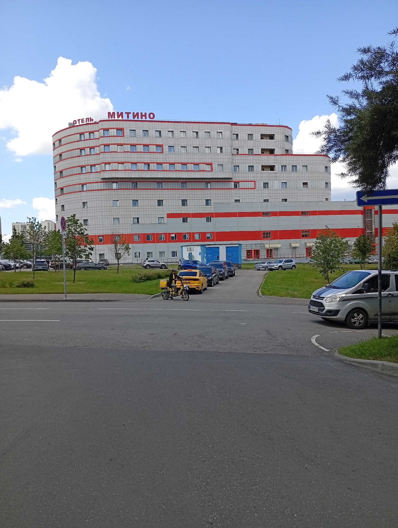 The image shows a multi-story hotel building with red and white horizontal stripes and red window frames, bearing the text "ОТЕЛЬ МИТИНО" in red lettering. In front of the hotel, a yellow taxi is parked next to a dark-colored car. A person is riding a yellow three-wheeled scooter with a cargo box. Several other cars are parked in a lot to the left of the hotel. A paved road and a grassy area are visible in the foreground, with a silver minivan turning right on the road. The sky is blue with white clouds.