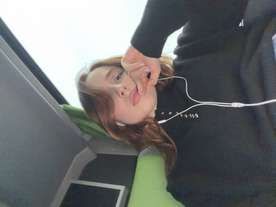 A young female, with shoulder-length reddish-brown hair parted on the left, is depicted from the chest up, taking a selfie inside what appears to be a passenger vehicle, likely a bus or coach, in Moineville, France. She wears a black ribbed turtleneck sweater with silver-colored ring fasteners on the collar and white wired earbuds in both ears, the cable extending down her chest. Her left hand is raised, with fingers slightly curled, thumb and index finger touching her lower lip area. A ring is visible on her left ring finger, and her fingernails are painted a light shade. Her lips are slightly parted, revealing a portion of her tongue, and she is looking directly at the camera. In the background, to the left, a portion of a green upholstered seat and a rectangular screen are visible. To the right, a large window shows a bright, overcast sky. A green curtain or fabric element is partially visible near the window's top left edge. The overhead ceiling panel is gray.