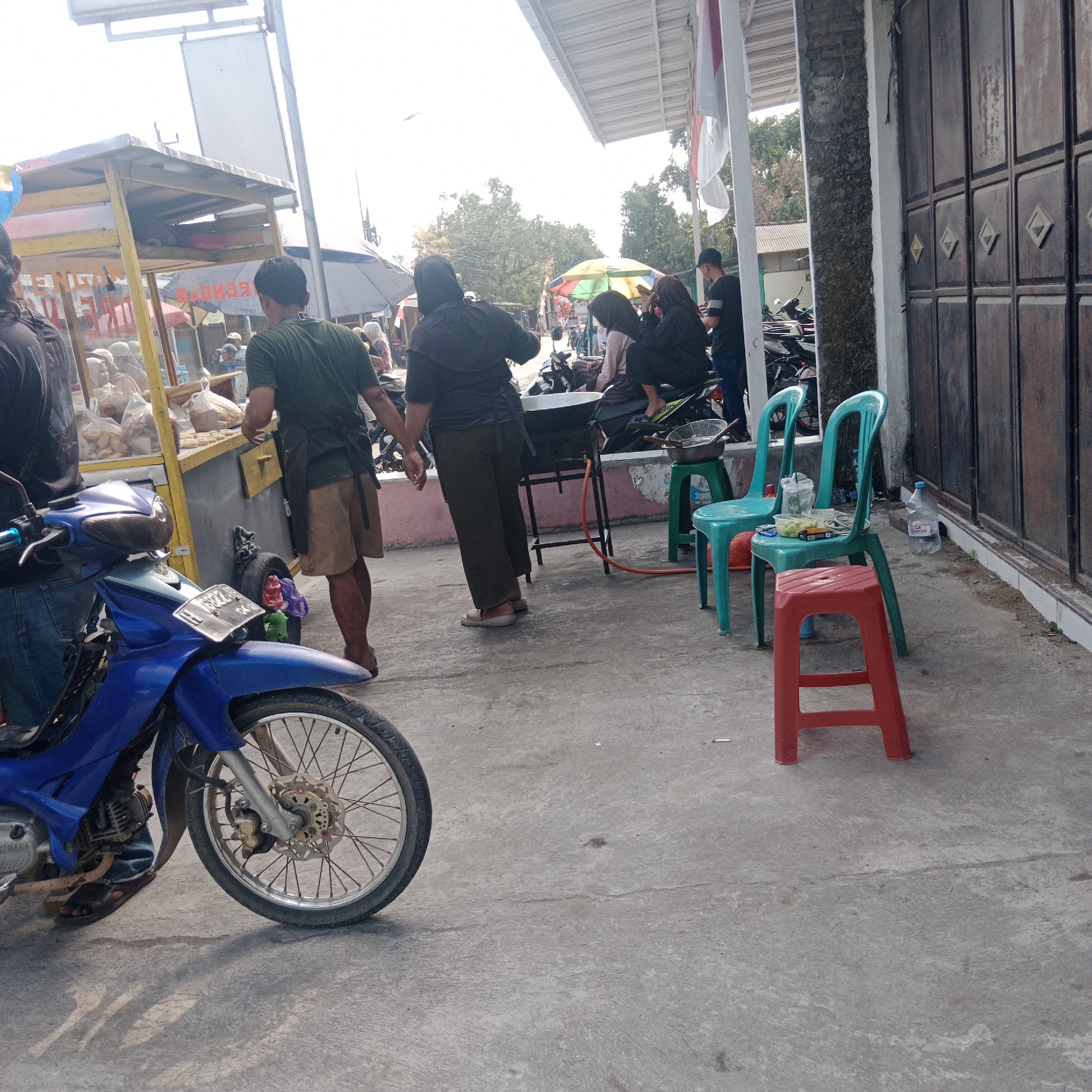 In Tegalsari, Indonesia, an outdoor scene unfolds with a food stall and surrounding activity. The stall, featuring a yellow and white structure with various food items displayed in plastic bags, is the central focus. A vendor in a green t-shirt and brown shorts stands behind the stall, appearing to serve customers or prepare food. Several individuals are gathered around the stall, some in dark clothing, including a person in a black headscarf. A blue motorcycle is parked in the foreground, its front wheel prominent. To the right of the food stall, three plastic chairs are arranged: two teal chairs and a red stool. A large plastic water bottle sits near the chairs. In the background, there are more motorcycles, trees, and a building with a dark brown metal door. The ground is paved with concrete, and the scene is illuminated by natural daylight.