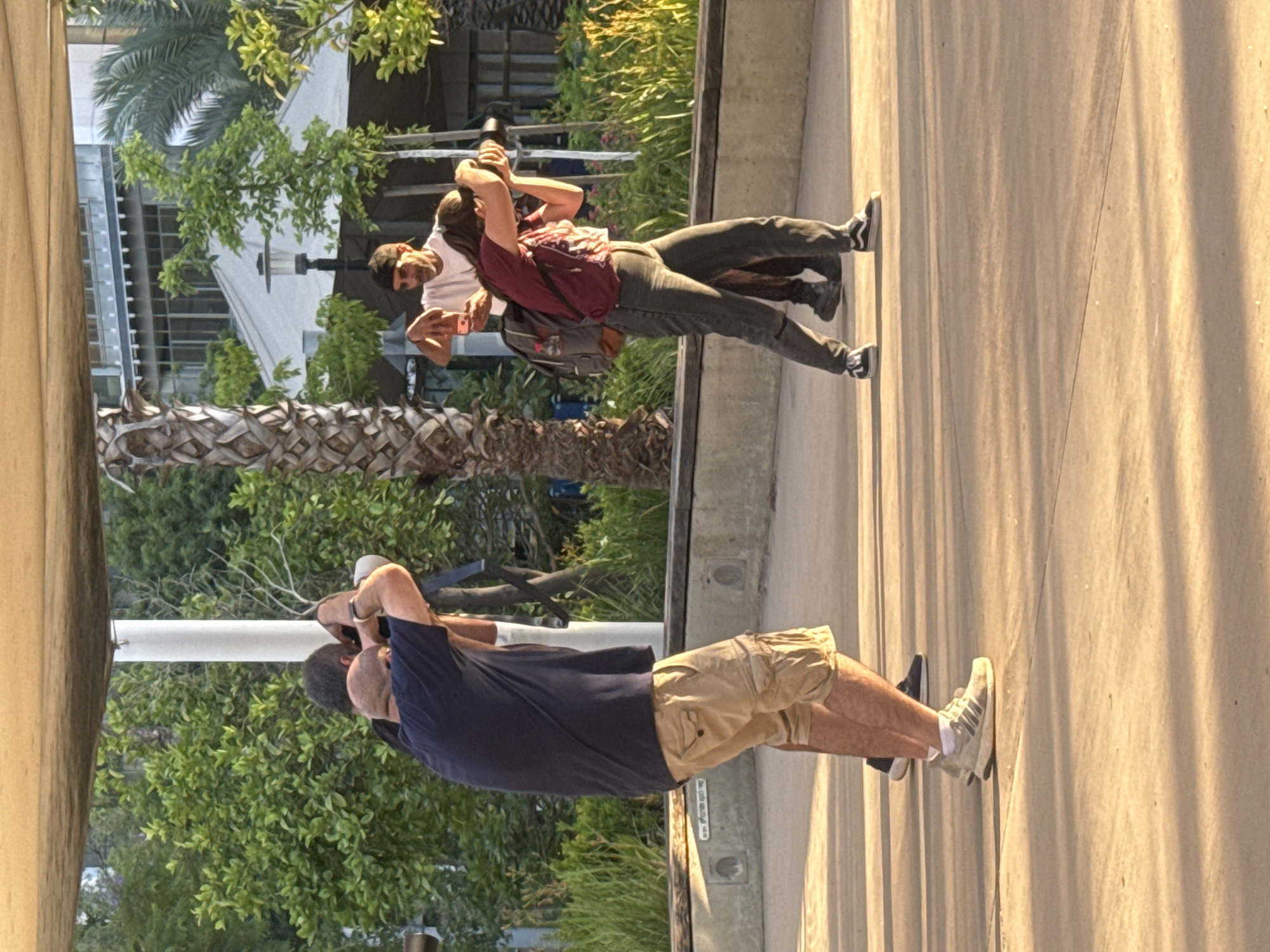Three individuals are captured in an outdoor setting in Tel-Aviv, Israel, engrossed in photography under warm, directional sunlight. The image, when oriented correctly, shows a concrete pathway on the right side, with a lush green backdrop of trees, including prominent palm trees, and modern buildings on the left.
In the foreground, closer to the viewer and towards the right, a man with a beard is visible. He wears a navy t-shirt, khaki cargo shorts, and light-colored sneakers, holding a professional camera to his eye, seemingly focused on a subject to his left. Slightly behind him and more centrally positioned, a woman in a dark red long-sleeved shirt, dark pants, and dark sneakers also holds a camera to her eye, mirroring his action. She has a backpack slung over her shoulders. Further back and to the left of the woman, another man in a white t-shirt, dark pants, and dark sneakers holds a smartphone in front of him, possibly taking a photo or recording.
The strong, golden light suggests either early morning or late afternoon, creating long, distinct shadows on the concrete path. The background features dense green foliage, including several large palm trees with rough, textured trunks, framing contemporary buildings with expansive windows. The scene conveys a sense of focused activity, with all three subjects actively engaged in capturing images of their surroundings. No visible text is present in the image.