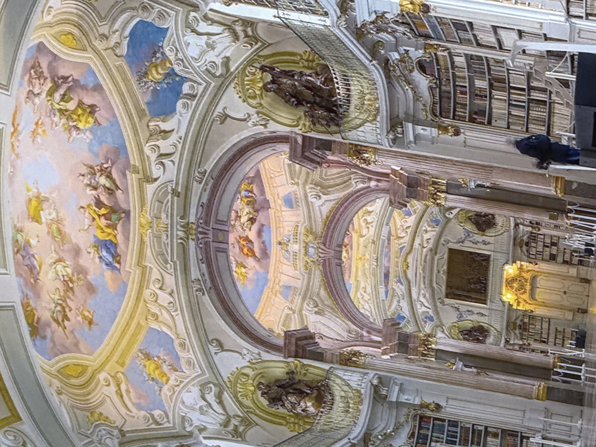 The image shows a grand, ornate library interior with towering bookshelves lining the walls. The ceiling is elaborately decorated with a fresco of angelic figures in a sky setting. Arches and decorative moldings are visible throughout the space, with gilded accents and large statues placed on balconies. A person in a dark jacket is seen standing near a bookshelf, possibly browsing or organizing. The overall impression is one of opulence and historical grandeur. There is no information in the image to suggest a location in Simferopol, Ukraine; the scene appears to be a European Baroque or Rococo style library.