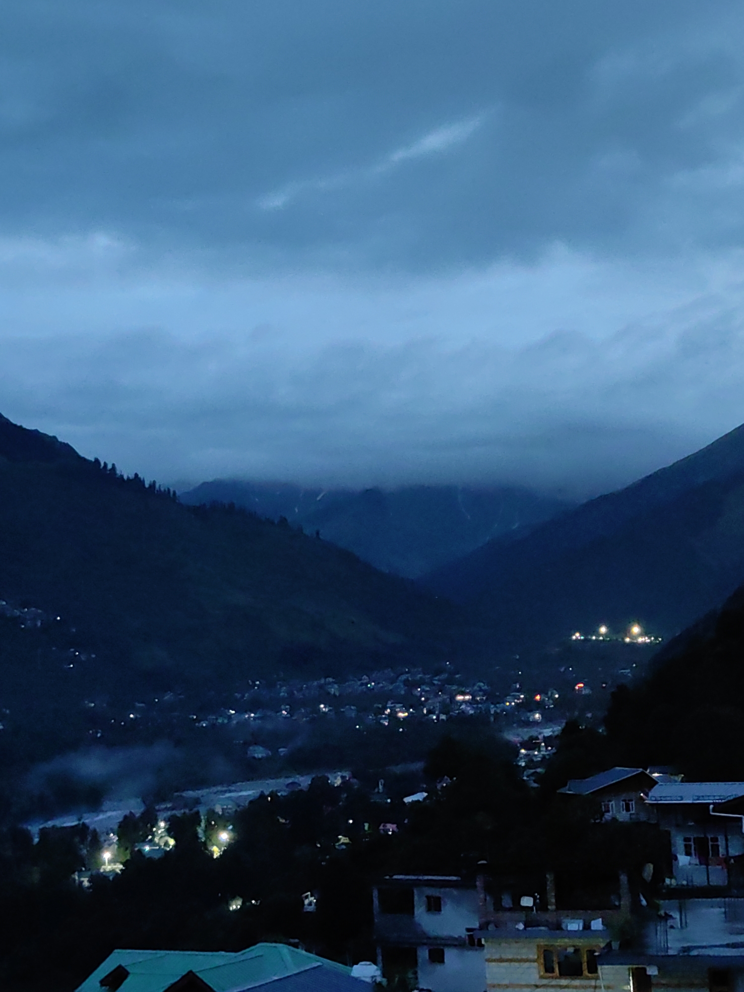 The image depicts a twilight or night scene in the mountainous region of Vashisht, India. The sky is overcast with dark, cloudy formations. In the foreground, the dark silhouette of a house with a teal-colored roof is visible. Beyond this, a village or town is spread out across a hillside, illuminated by scattered lights. Trees are interspersed throughout the settlement. Further in the background, the imposing shapes of mountains rise, with some peaks obscured by clouds. The overall atmosphere is dark and serene, with the village lights offering a sense of human presence amidst the natural landscape.