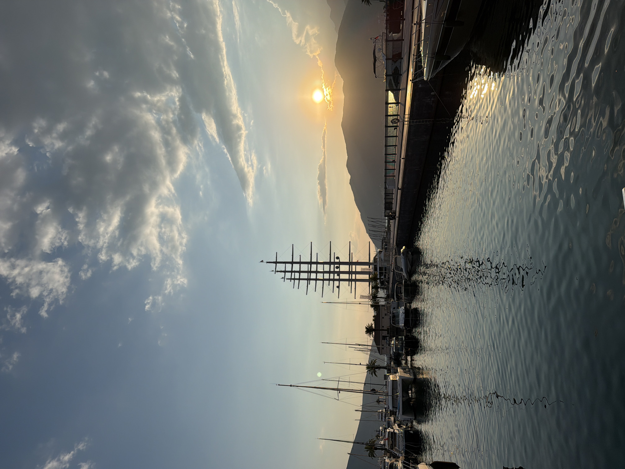 The image depicts the marina in Tivat, Montenegro, at sunset. The sky is a gradient of pale blue and orange, with scattered clouds. The sun, a bright orb, is partially obscured by clouds and mountains in the distance, casting a warm glow across the scene. A row of sailboats and yachts are moored in the foreground, their reflections shimmering on the calm water. A large, multi-masted sailing vessel stands prominently among them, its dark silhouette contrasting with the bright sky. The marina features a pier with railings, and in the background, mountains form a silhouetted backdrop. The overall impression is one of tranquility and maritime elegance.