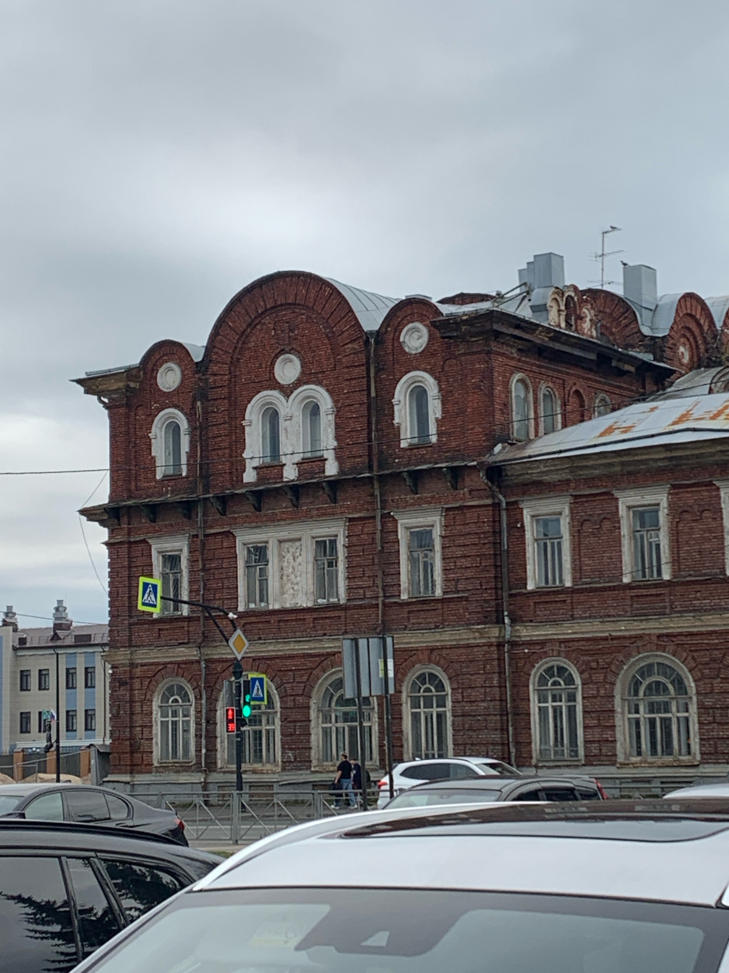 The image captures a street view in Arkhangelsk, Russia, on an overcast day. In the foreground, several cars are parked or in motion, obscuring the lower part of the scene. Dominating the midground is a large, multi-story brick building with an ornate, arched facade and numerous windows. Some of the windows feature distinctive arched tops, framed by white detailing. The building's architecture suggests it is an older structure.
A traffic light with a green pedestrian signal is visible, indicating it is safe for pedestrians to cross. A pedestrian crossing sign is also present. In the background, another building with a more modern facade, featuring horizontal blue and white stripes, is partially visible. The sky is a uniform gray, typical of cloudy weather, contributing to the muted overall lighting of the scene.