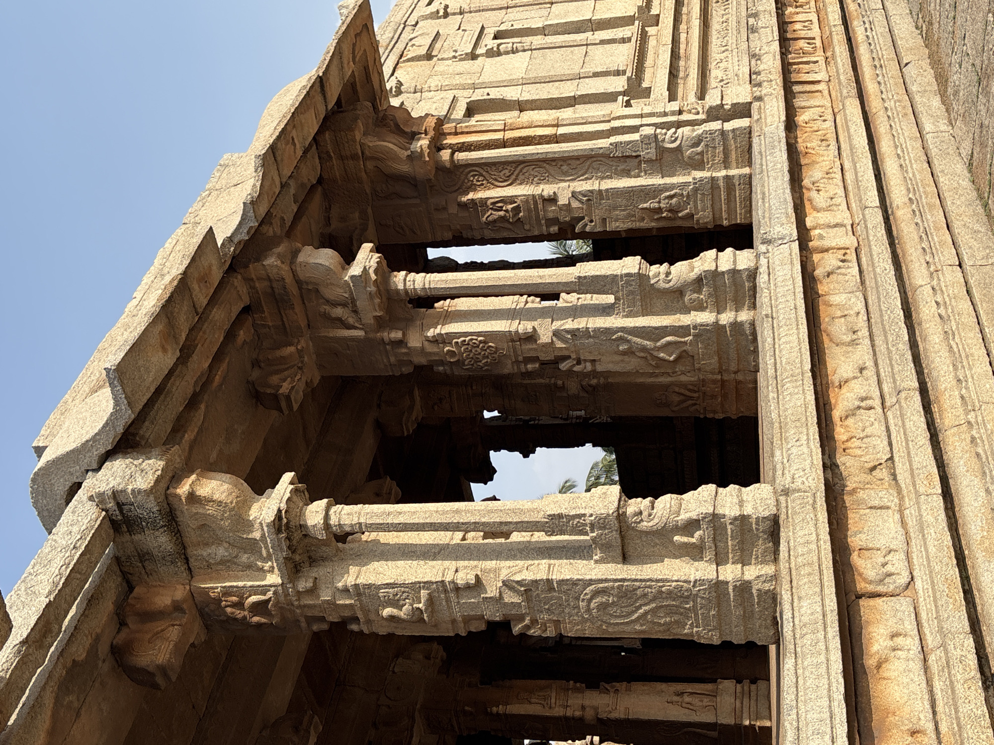This is a low-angle shot of the intricate stonework on an ancient building in Powerhouse Colony, India. The building appears to be a temple or a similar religious structure, judging by the carvings and architectural style. The stonework features stacked layers of carved columns and beams, creating a tiered effect. There are openings between the layers, providing glimpses of the sky and some foliage. The overall color palette is warm, dominated by the beige and tan tones of the stone under the bright sunlight. The sky is a clear, pale blue. The perspective is tilted upwards, emphasizing the height and complexity of the structure.