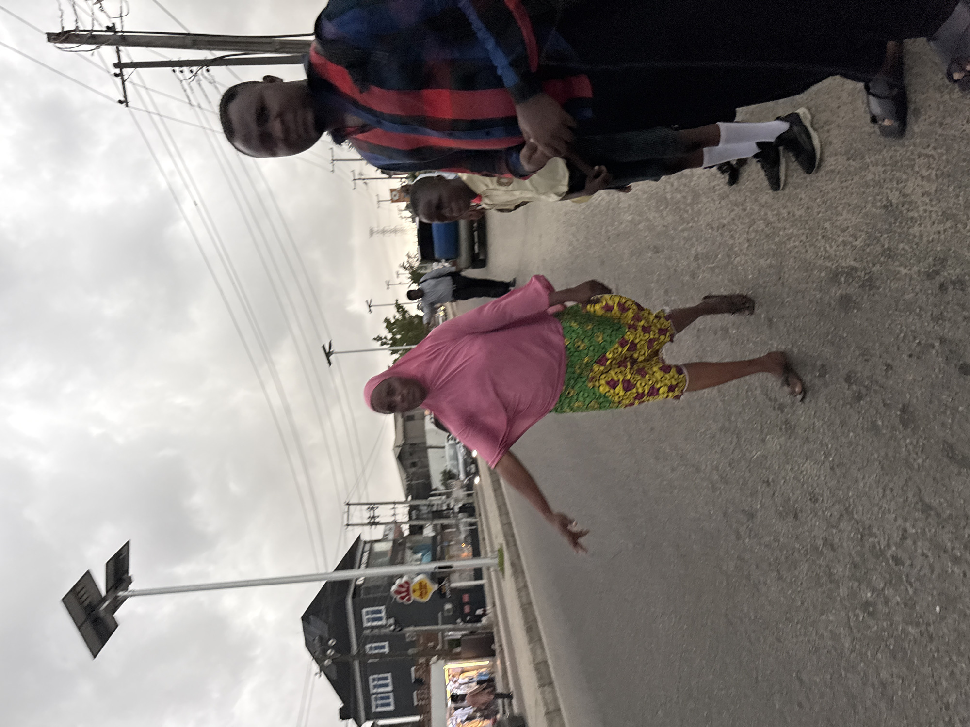 The image captures a street scene in Ado, Nigeria, on an overcast day, with several people walking and standing. The perspective is tilted, making the ground appear as a slanted surface.
In the prominent foreground, to the left, a man in a red, blue, and black plaid long-sleeved shirt and dark trousers looks towards the viewer. He stands near a child, likely male, wearing a light-colored (possibly cream or yellow) long-sleeved shirt, dark shorts or trousers, white socks, and dark sneakers. This child is positioned slightly behind and to the right of the man in plaid. Further back, another individual, partially obscured, is visible between the first man and child.
To the right in the mid-ground, a woman is captured in motion, appearing to walk or gesture. She wears a striking pink long-sleeved top and a vibrantly patterned skirt featuring green, yellow, and red designs. Her feet are bare. Behind her, another man in a light-colored shirt and dark pants stands near a blue vehicle.
The setting is a paved road with a sidewalk on the left. Utility poles with numerous overhead wires stretch across the cloudy sky. Buildings, some appearing commercial or residential, line the street. One building features a red and yellow logo that resembles a chicken head, possibly indicating a restaurant or fast-food establishment. Other signs on buildings and street furniture are present but illegible due to distance and blur. The overall impression is that of a bustling local street in daytime, despite the cloudy conditions.