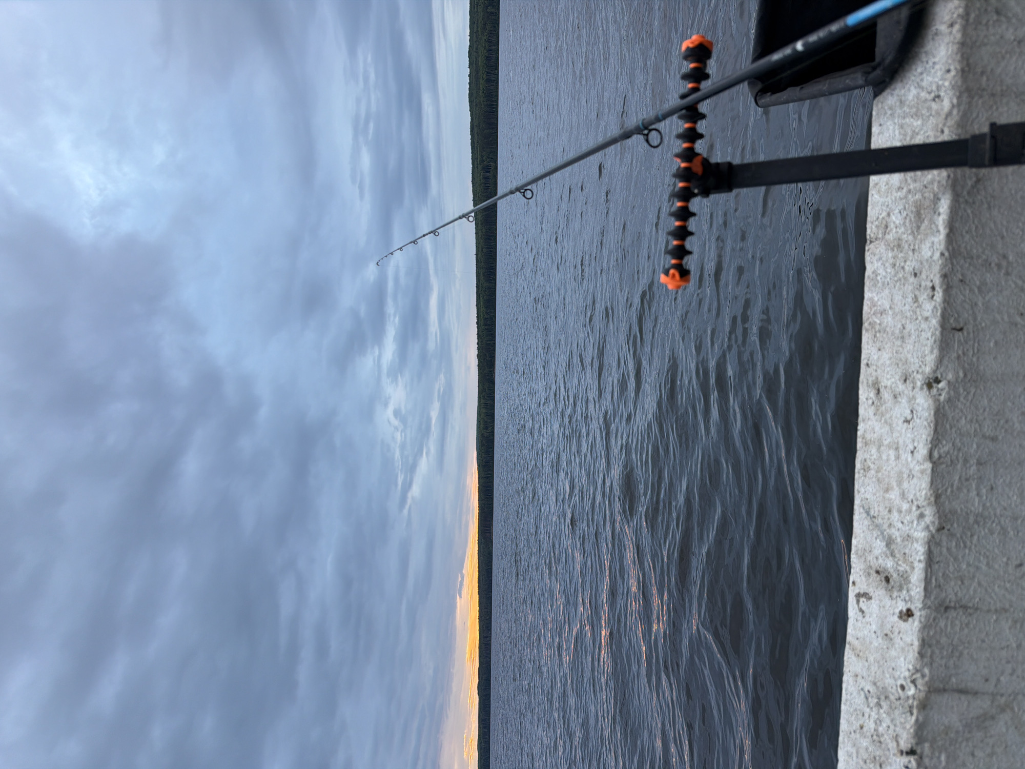 The image depicts a fishing scene at dusk or dawn over a large body of water in Chaikovsky, Russia. In the foreground, a rough-textured, light grey concrete structure extends across the bottom of the frame. Resting on this structure is a black fishing rod holder with distinct orange, ribbed accents, supporting a dark grey fishing rod that angles upwards and to the right, displaying multiple line guides. A dark container or bag is partially visible to the right of the rod holder. The water, a deep blue-grey, exhibits moderate ripples, with a faint orange reflection from the sun visible on its surface towards the left. A dense, dark green treeline forms a distinct horizontal band on the distant horizon. Above the treeline, a narrow strip of orange light indicates the setting or rising sun, partially obscured by dark, textured grey clouds that dominate the upper sky. No individuals are visible.