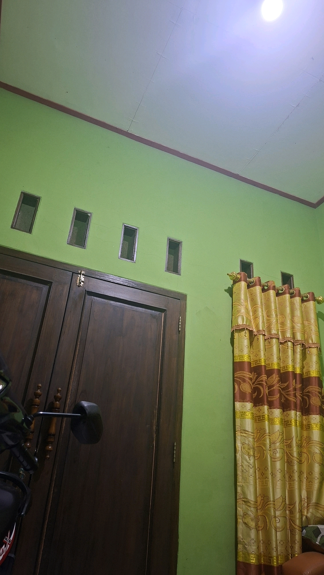 The image captures an interior scene of a room. The walls are painted a vibrant green, and the ceiling is white with visible seams and a bright light source at the top right. A thin, dark brown trim runs along the intersection of the walls and ceiling.
On the left side of the image, there is a large, dark brown wooden double door. The doors appear to be made of solid wood and have a traditional design with visible grain. A metallic hinge is visible on the top of the right door, and a decorative wooden knob with carvings is present on the left door. A black motorcycle mirror with a reflective surface is mounted on a metal arm extending from the left side of the door frame, partially obscuring the left door.
Above the doors, there are four small, rectangular openings in the green wall, spaced evenly apart. These openings are framed with a dark material and appear to be ventilation or decorative elements.
On the right side of the image, a set of decorative curtains hangs. The curtains are a combination of golden yellow and dark brown, with an ornate pattern featuring swirling motifs and geometric shapes. The curtains are drawn, revealing three more small, rectangular openings similar to those above the door, which appear to be placed at window height. The overall impression is of a residential interior, possibly in Sranten, Indonesia, given the context.
