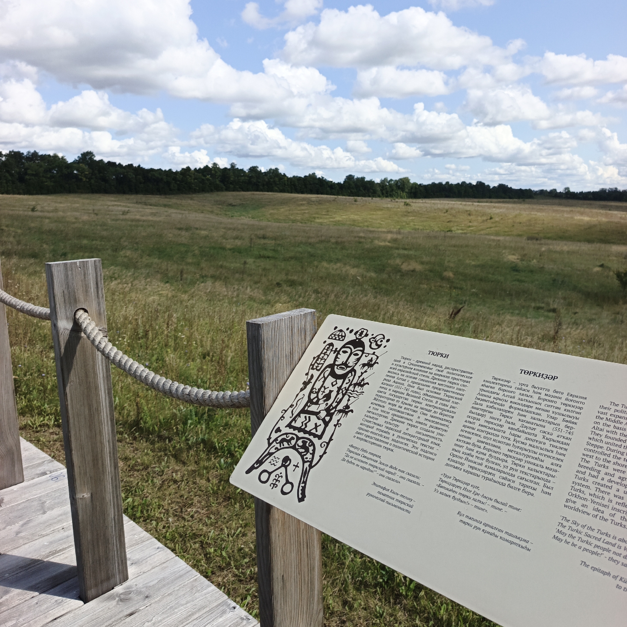 The image shows an outdoor informational display in Verkhnie Termy, Russia, situated within a natural landscape. The display consists of a wooden post and rope barrier, with a sign featuring text and illustrations in both Cyrillic and Latin scripts. The sign details information about the Turkic people and their historical context, including illustrations of what appear to be ancient artifacts or symbols. The background depicts a grassy field bordered by a dense line of trees under a partly cloudy sky. The overall scene suggests an archaeological or historical site open to the public, emphasizing the cultural heritage of the region.