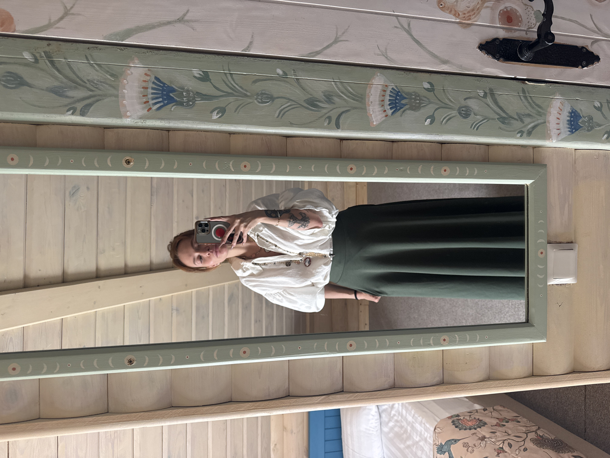 A woman is taking a mirror selfie in a room with wooden walls. She is wearing a white voluminous blouse and a long dark green skirt. She has several necklaces on and multiple tattoos visible on her arms. A phone is visible in her hand, held up to take the photo. The mirror has a decorative frame painted with a folk art motif. To the left of the mirror, a bed with patterned bedding is visible. To the right of the mirror, a wooden door with painted floral designs and a black door handle is present. The room appears to be part of a guesthouse or hotel, possibly in Suzdal, Russia, given the style of the wooden architecture and folk art decorations.
