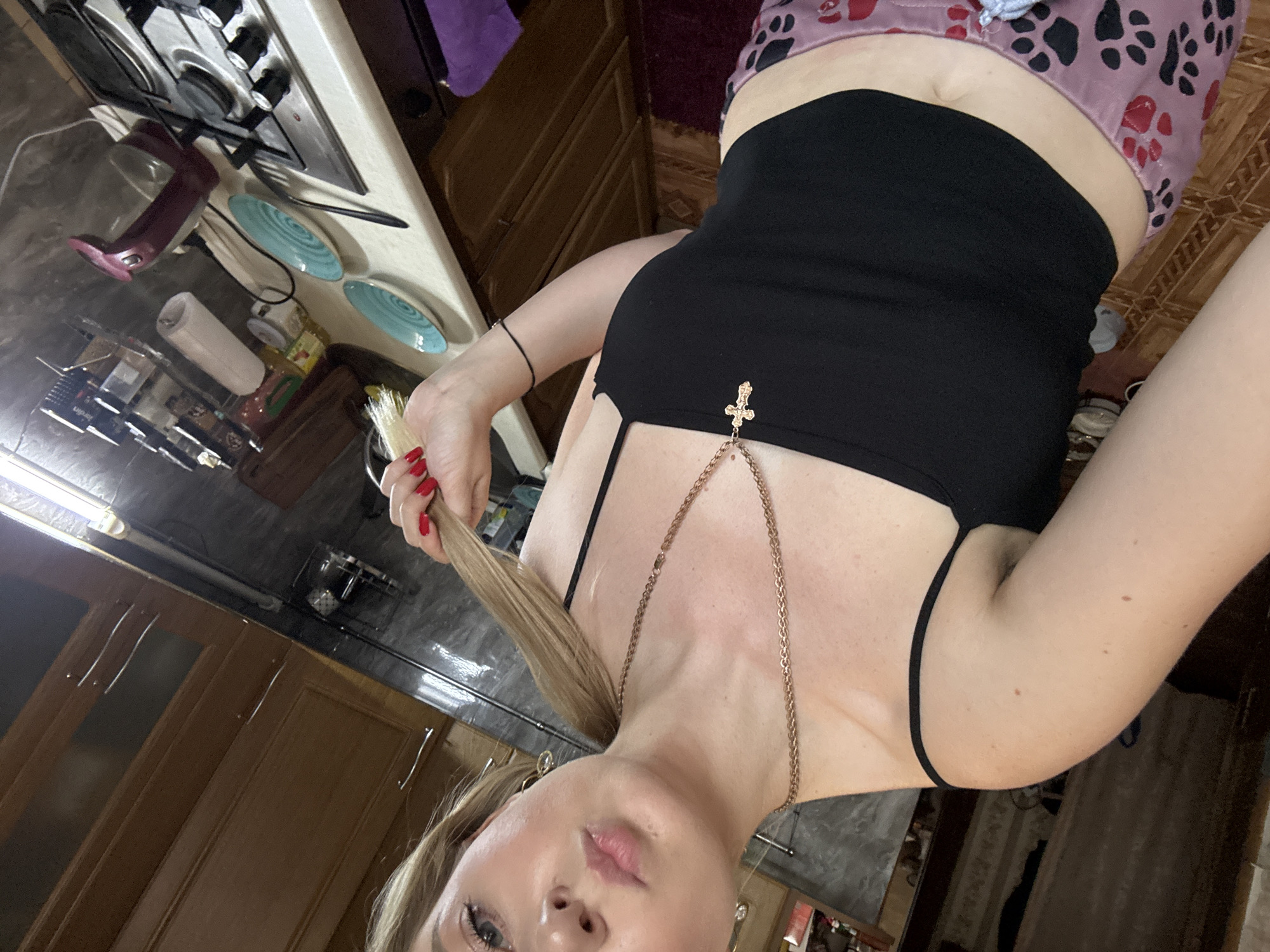 A young woman with blonde hair pulled into a ponytail, wearing a black crop top and pink pajamas with paw prints, poses for a selfie in a kitchen. She is holding a strand of her hair and her right hand is near her face. A gold necklace with a cross pendant hangs around her neck. The kitchen has wooden cabinets, a stovetop, and various items on the counter, including plates and a kettle. The scene appears to be indoors.