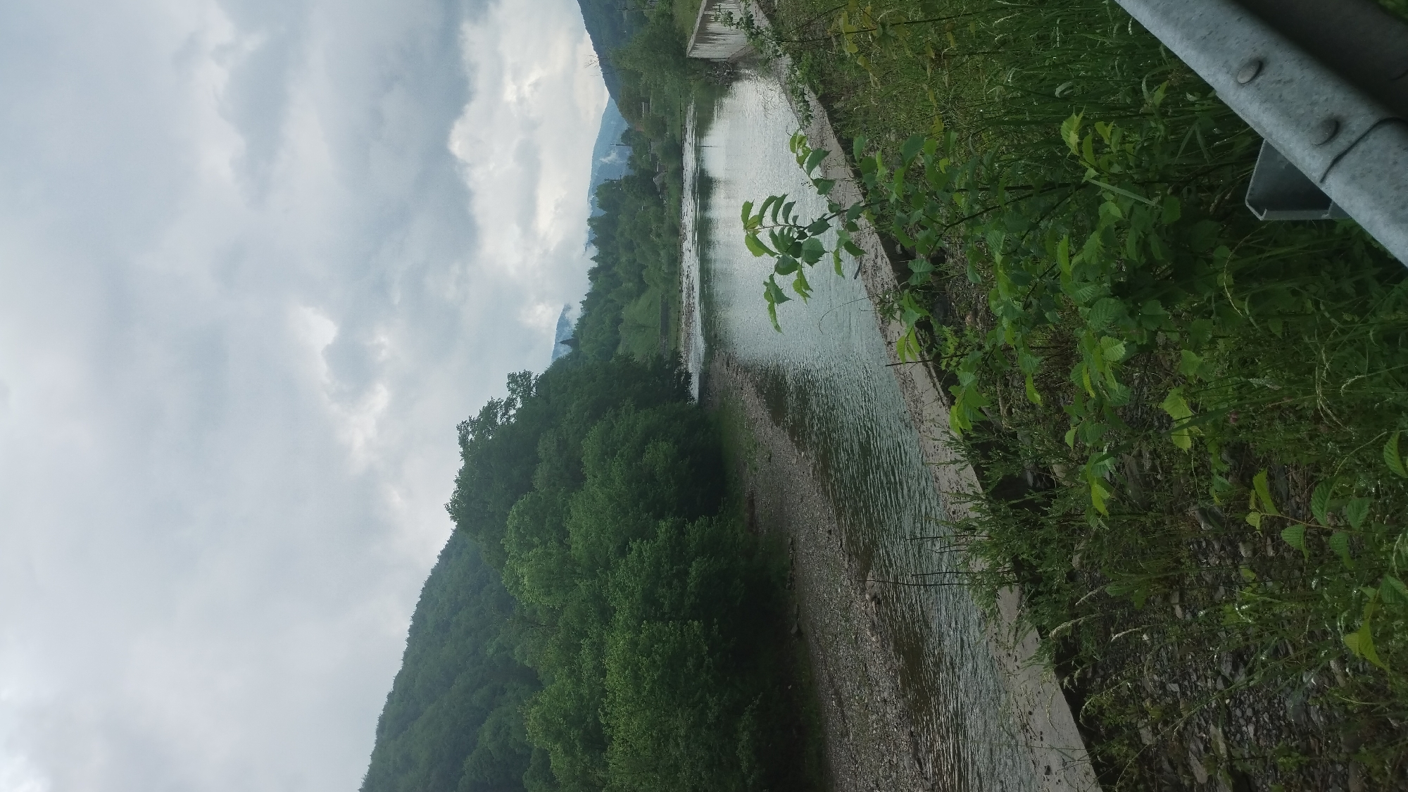 The image shows a river flowing through a landscape in Trostianets, Ukraine. The riverbanks are lined with lush green vegetation and trees, and a mountain range is visible in the background. A concrete barrier and metal guardrail run along the riverbank. The sky is overcast with heavy clouds.
