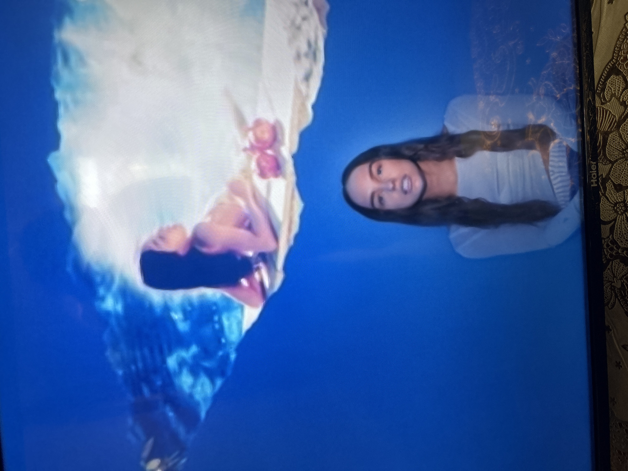 This media file is a photograph of a television screen displaying a surreal image. The image features two women. One woman appears to be submerged in water or a similar fluid, with her body partially obscured by billowing white fabric and pink flowers. The water or fluid is a vibrant blue. The second woman is visible on the right side of the screen, looking towards the camera. She has long, dark hair and is wearing a light-colored top. The overall scene is abstract and dreamlike, with a strong contrast between the deep blue background and the lighter elements of the image. There are no clear indicators of time of day, weather, or specific location cues, beyond the provided context of Gelendzhik, Russia. The emotions conveyed are ambiguous, leaning towards a sense of mystery or fantasy. A visible text label on the television screen reads "Haier."