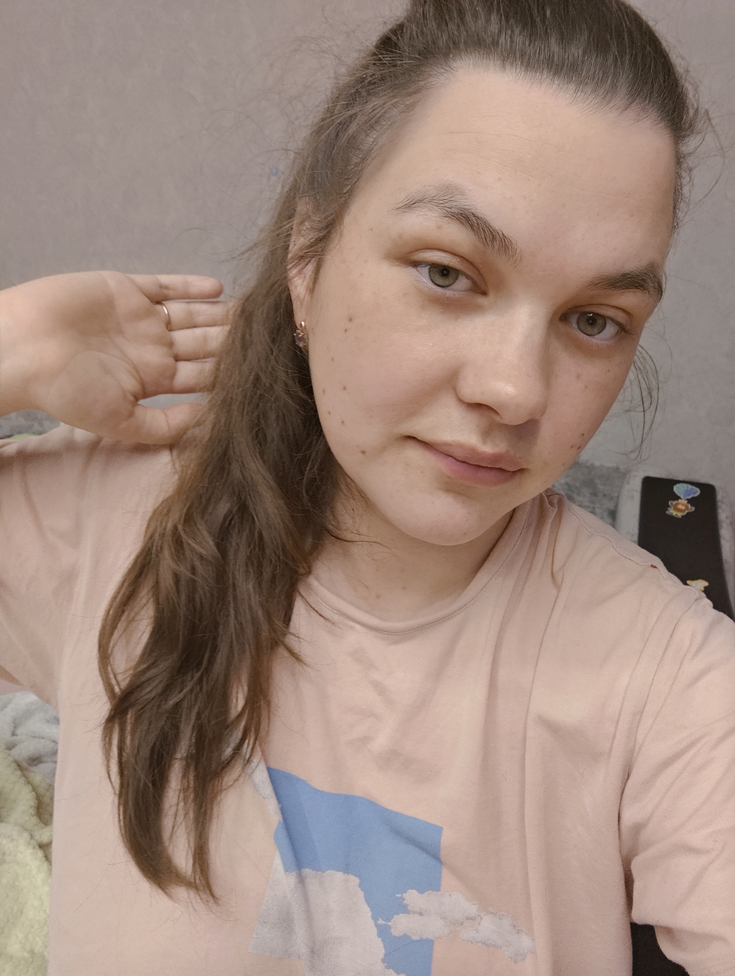 A young woman with light skin, light brown hair pulled back in a ponytail, and freckles poses for a close-up selfie indoors. She wears a light pink t-shirt with a blue and white design. Her left hand is raised near her head. A ring is visible on the ring finger of her left hand. The background includes a light-colored wall and a glimpse of a phone case or device with stickers on a dark surface. The location is Chaikovsky, Russia.
