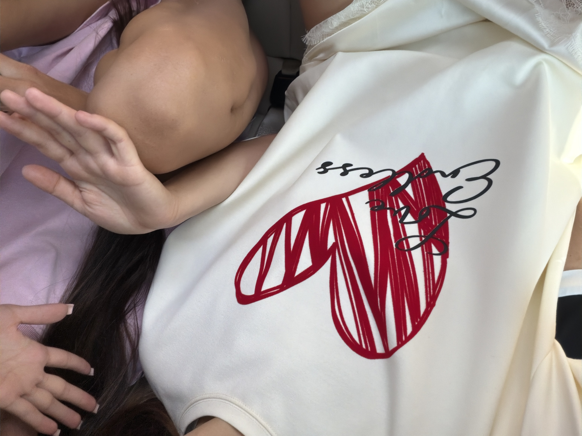The image is a close-up, high-angle shot of a person wearing a white t-shirt with a large red heart graphic and black cursive writing. The writing appears to say "Lovely" or "Love," with a stylized "s" at the end. The t-shirt is positioned across the frame, with the graphic prominent. To the left of the t-shirt, a person's hands with manicured nails are seen resting on dark hair. To the right of the t-shirt, a portion of another person's knee and upper thigh is visible, suggesting they are sitting or lying down. The background is blurred, with hints of pink fabric and a grey upholstered surface, possibly a car seat. The lighting is soft and diffused. There is no visible indication of Arpașu de Jos, Romania.