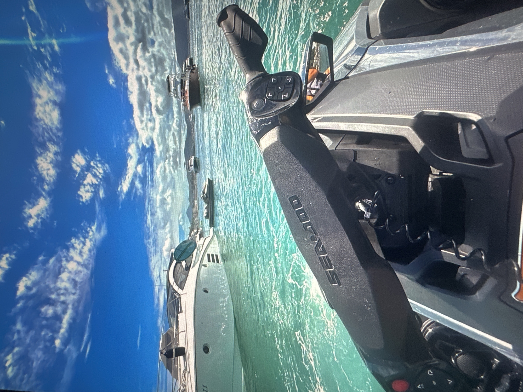 The image depicts a personal watercraft, a Sea-Doo, positioned on turquoise water under a vibrant blue sky with scattered clouds. The handlebars of the watercraft are prominent in the foreground, displaying control buttons and a Sea-Doo logo. In the background, several boats, including a large white yacht and a smaller dark-colored boat, are anchored in the water. The shoreline, characterized by hills and buildings, is visible in the distance. The scene suggests a recreational outing on a sunny day in a coastal area. There are no people clearly visible in the image.