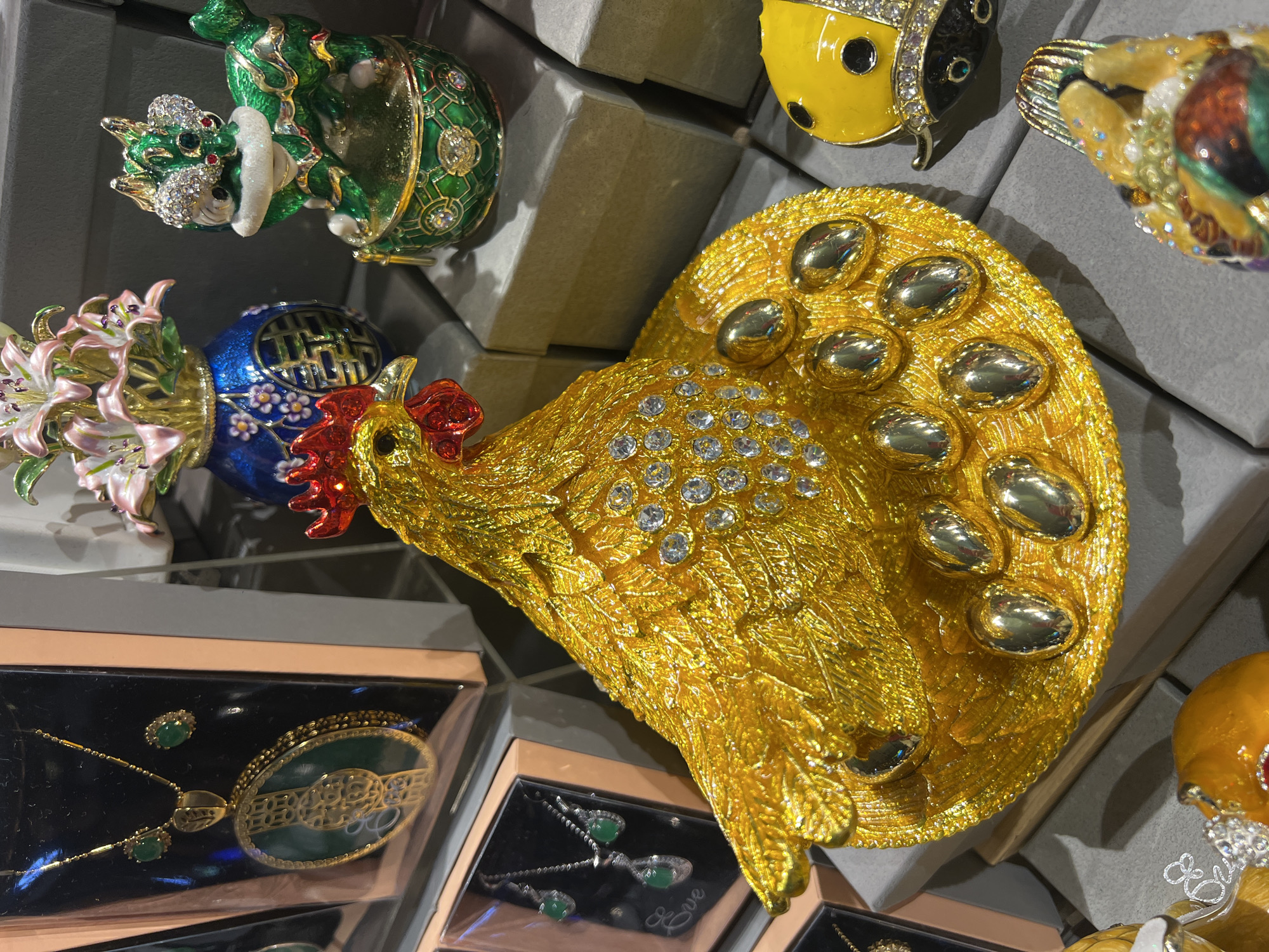 This close-up image captures a vibrant display of ornate, jewel-encrusted decorative objects and jewelry, likely within a retail setting such as a souvenir or gift shop in Kuta, Indonesia.
The central and most prominent item is a large, golden rooster figure. Its body and tail feathers are highly textured with a rich gold finish, adorned with numerous clear rhinestones and larger, smooth gold oval accents on its tail. The rooster's comb and wattle are a bright red.
Surrounding the rooster are other elaborate trinket boxes or decorative figurines. To the upper left, a green creature, possibly a dragon or turtle, is intricately patterned and studded with clear rhinestones and gold details. Below it, a blue vase with gold patterns, possibly including a Chinese character resembling "福" (fú) for good fortune, holds a bouquet of pink lily-like flowers with sparkling accents. To the upper right, part of a yellow ladybug-shaped item with black spots and rhinestones is visible, alongside another colorful, possibly bird-shaped item. A small, orange, rounded object is partially seen at the bottom right.
Beneath these larger items, several jewelry sets are displayed in black cases. On the bottom left, a necklace with a large, oval green pendant featuring intricate gold filigree and matching stud earrings is shown. In the center bottom, another jewelry set features a necklace and earrings with teardrop-shaped green stones, next to which the word "Eve" is visible on the case. The items are arranged on and around simple grey display boxes. The lighting suggests an indoor, brightly lit environment.