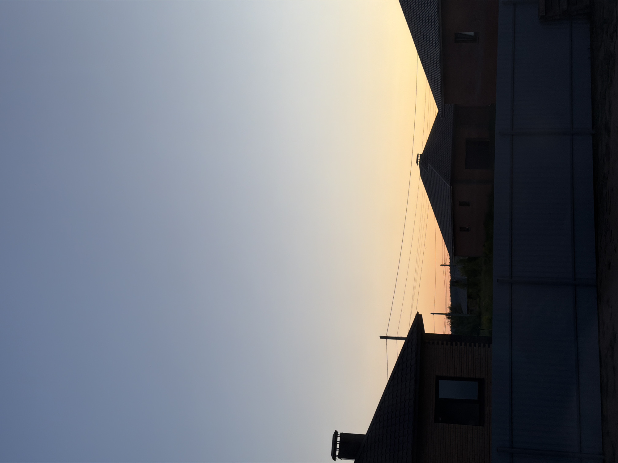 The image depicts a dusk scene in the city of Chaikovsky, Russia. Three brick houses with dark tiled roofs are visible. The house on the left has a prominent chimney. Power lines are strung between poles in the middle ground. In the foreground, a dark gray metal fence runs horizontally across the frame, obscuring the lower portions of the houses and the ground. The sky transitions from a pale blue at the top to a warm orange and yellow hue near the horizon, indicative of sunset. There are no people or animals visible in the image. The environment appears to be a residential area.