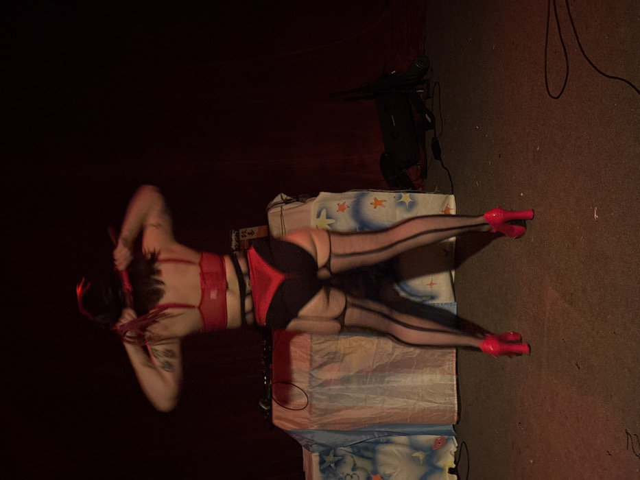 A person wearing red lingerie, black shorts with red details, black stockings, and pink high heels, is lying on a light-colored surface. The surface is printed with blue and yellow shapes. A black object with wires is behind the surface. The background is dark. The person has tattoos on their arms and back. They are holding what appears to be black feathers behind their head.