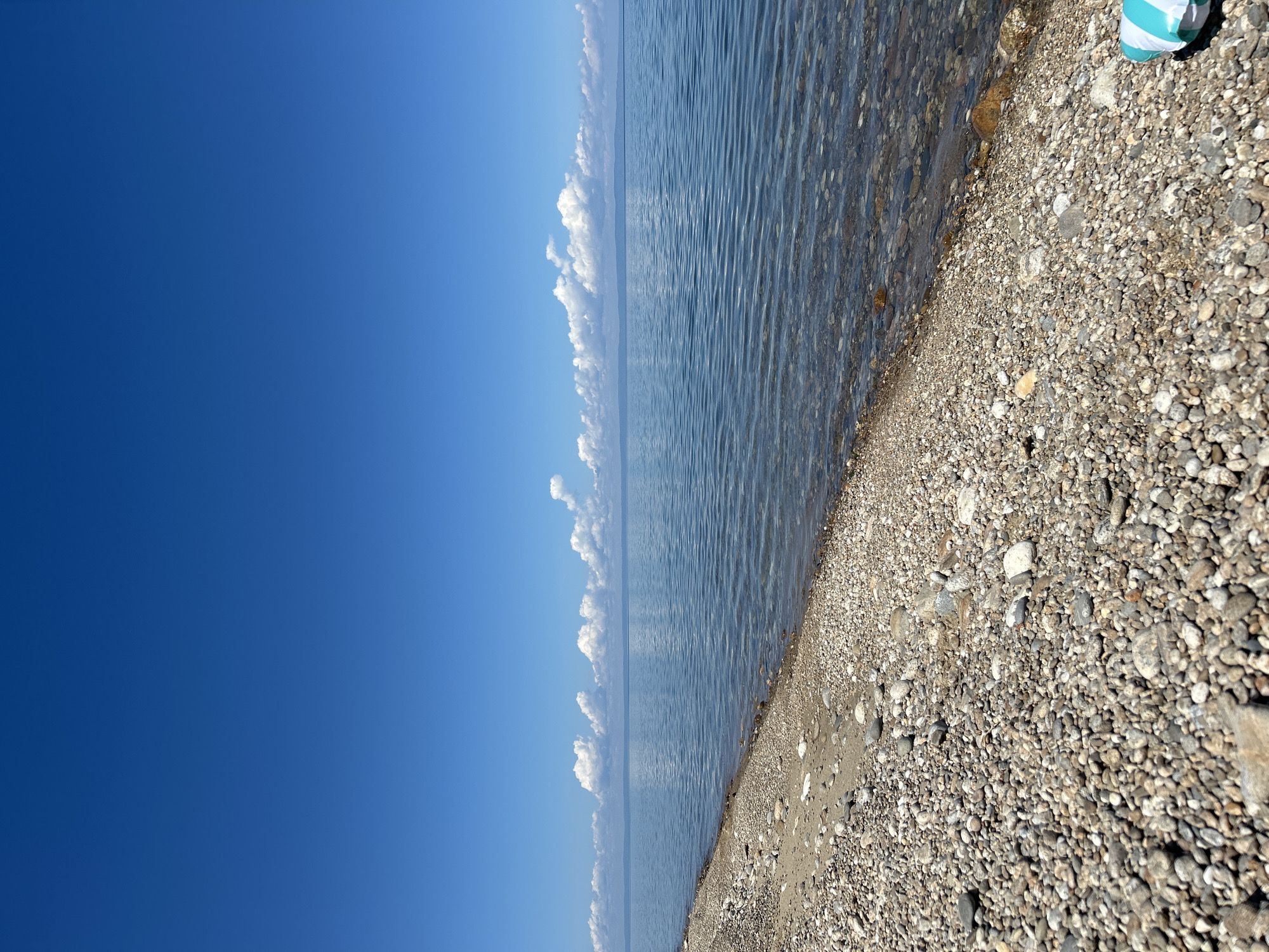 The image depicts a serene beach scene under a clear blue sky, with a vast expanse of calm water stretching to the horizon. The foreground is a pebbly beach, with smooth, varied-colored stones leading down to the water's edge. The water itself is shallow and clear, revealing more stones beneath the surface. A faint line of clouds dots the distant horizon, separating the water from what appears to be a landmass. A portion of a blue and white striped object, possibly a beach mat or float, is visible in the lower right corner of the frame, suggesting human presence or recent activity. The overall impression is one of tranquility and natural beauty. The scene is identified as being in the city of Tolbazikha, Russia.