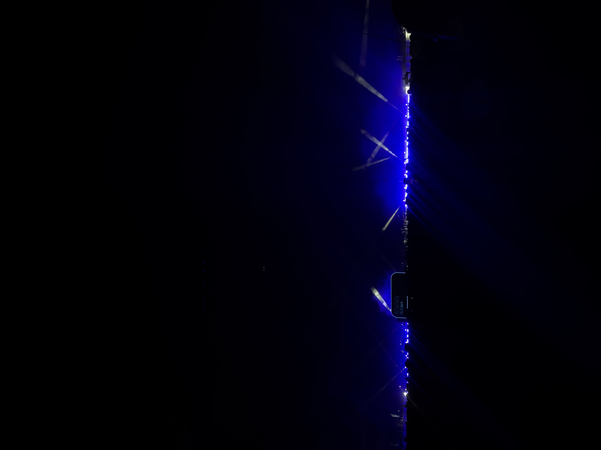The image depicts a dark scene, possibly at night, with blue lighting and beams. A vertical structure on the right side is illuminated with a string of blue lights. Light beams in a crisscross pattern are also visible. There's a sign with text that reads "LOMA," with other illegible text above. It's unclear if the location mentioned in the prompt is accurate based on the visual information available. Due to darkness and lighting conditions, details regarding the environment, people, subjects and actions are unclear.