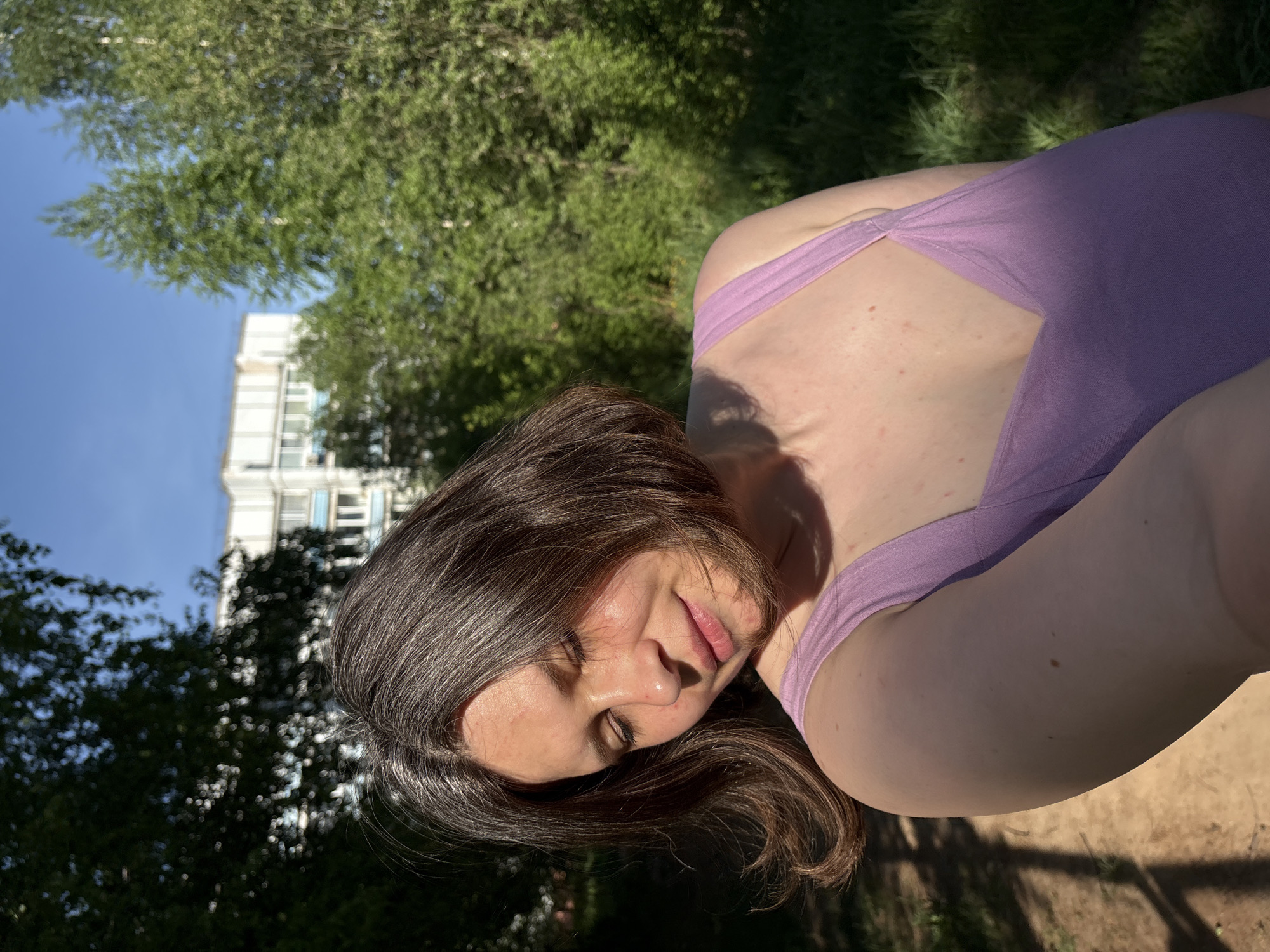 A woman wearing a purple tank top reclines in an outdoor setting in what is likely a park in Chaikovsky, Russia. The sun is shining brightly, casting shadows on the ground and on her face. Her dark hair with hints of grey is partially obscuring her face. In the background, a tall residential building is visible through the trees.