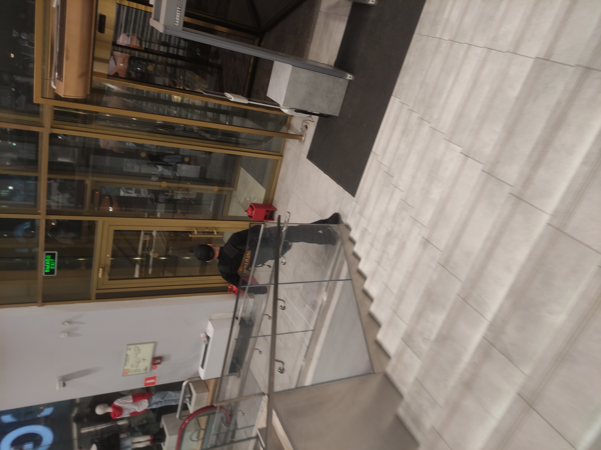 Here is a forensic description of the image:
**Location:** The image appears to be taken inside a commercial building, possibly a shopping mall, in Moscow, Russia.
**Environment:** The setting is modern, with a glass-walled storefront and staircase. The interior features tiled flooring and glass railing on the staircase.
**Objects:** Visible objects include a metal detector at the entrance, an exit sign in Russian, a fire safety diagram, and a mannequin.
**People:** A man dressed in black clothing is present on the stairway, and a mannequin is visible.
**Actions/Interactions:** The man appears to be walking up the staircase.
The scene appears to be a blend of security measures (metal detector) and retail display (mannequin) within a modern commercial setting.