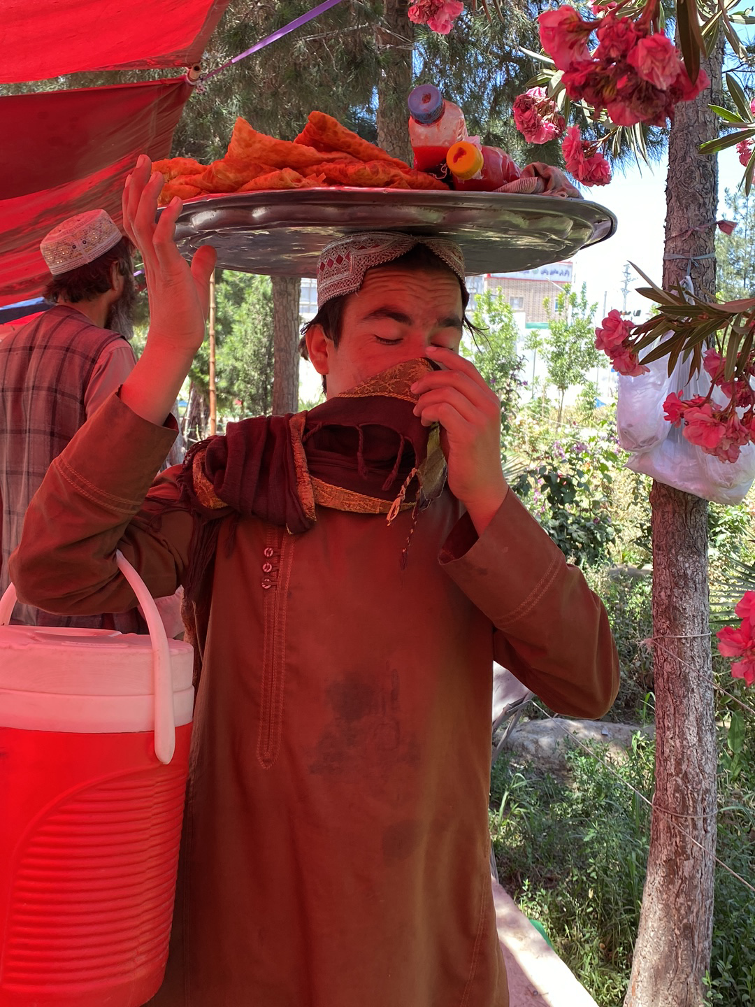 In a medium shot taken in the city of قندهار, Afghanistan, a young man with dark hair is seen carrying a large silver tray filled with pastries and a beverage bottle balanced on his head. He wears a white embroidered cap and a long-sleeved brown garment. His face is partially covered by a brown cloth. The man is positioned under a red awning, with a red cooler visible beside him. A person with a similar cap and a patterned shirt stands to the left, partially out of focus. In the background, there's greenery and a building. To the right of the subject, pink flowers hang from a tree trunk.
