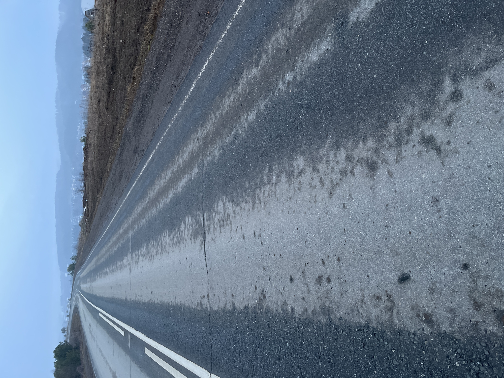 The image depicts an asphalt road in Фоки, Russia. The road surface is wet or damp, with varying degrees of wetness creating streaks and patches. A double white line runs along the left side of the road, indicating lane separation. To the left of the road is a grassy embankment with brown vegetation, possibly due to the time of year or lack of maintenance. Beyond the embankment, a hazy or foggy cityscape is visible in the distance, suggesting either inclement weather or atmospheric pollution. The overall scene is bleak and overcast. No people, vehicles, or other distinct objects are visible on the road or in the immediate vicinity. The road stretches into the distance, converging towards the horizon.