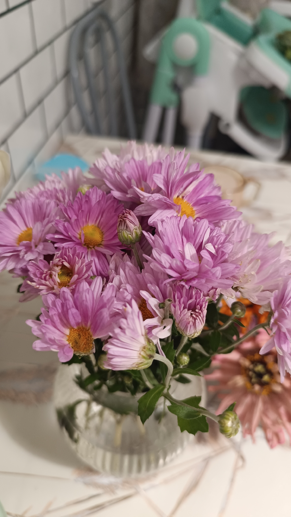 A close-up, vertical, eye-level shot focuses on a bouquet of vibrant pink chrysanthemums arranged in a clear, ribbed glass vase. The flowers, in various stages of bloom, display delicate petals and yellow centers. The background is softly blurred, revealing hints of a tiled backsplash, a high chair in muted green and white, and a patterned tabletop. The lighting appears natural, casting soft shadows and highlighting the texture of the petals. The scene is set indoors, likely in a domestic environment in Chaikovsky, Russia.