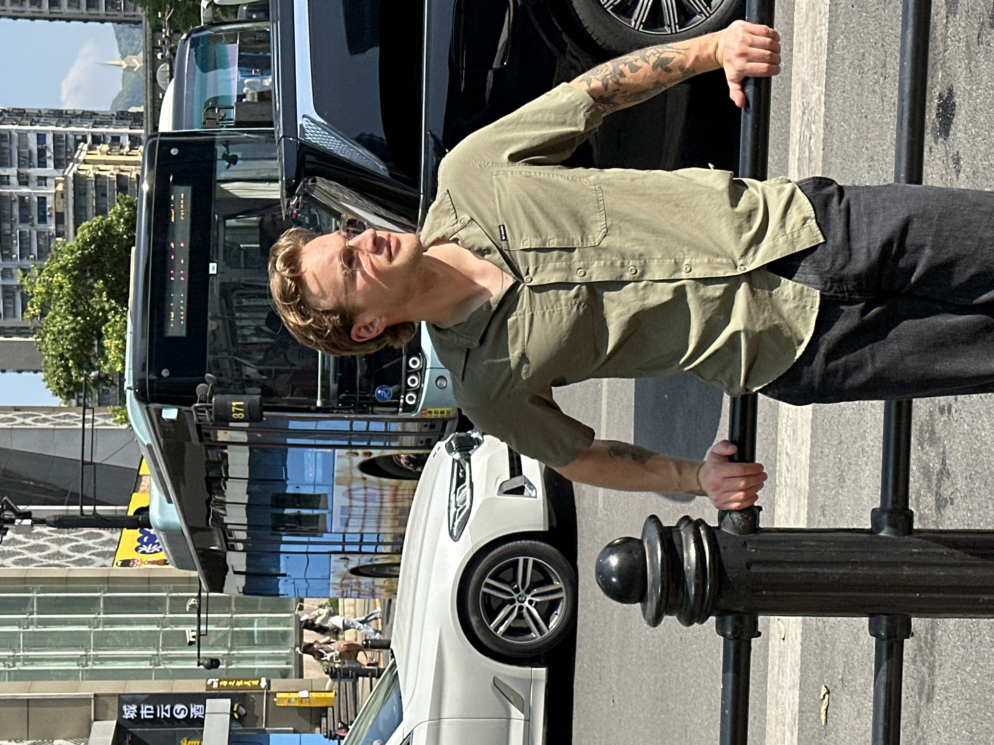 A man stands holding onto a black metal railing in what appears to be an urban area. The man is wearing a button-down shirt, dark pants, and glasses, and has tattoos on his arms. Behind him, there is a variety of vehicles on the road, including a black SUV, a bus, and a white BMW. In the background, tall buildings suggest the location is in the Yuzhong District of China.