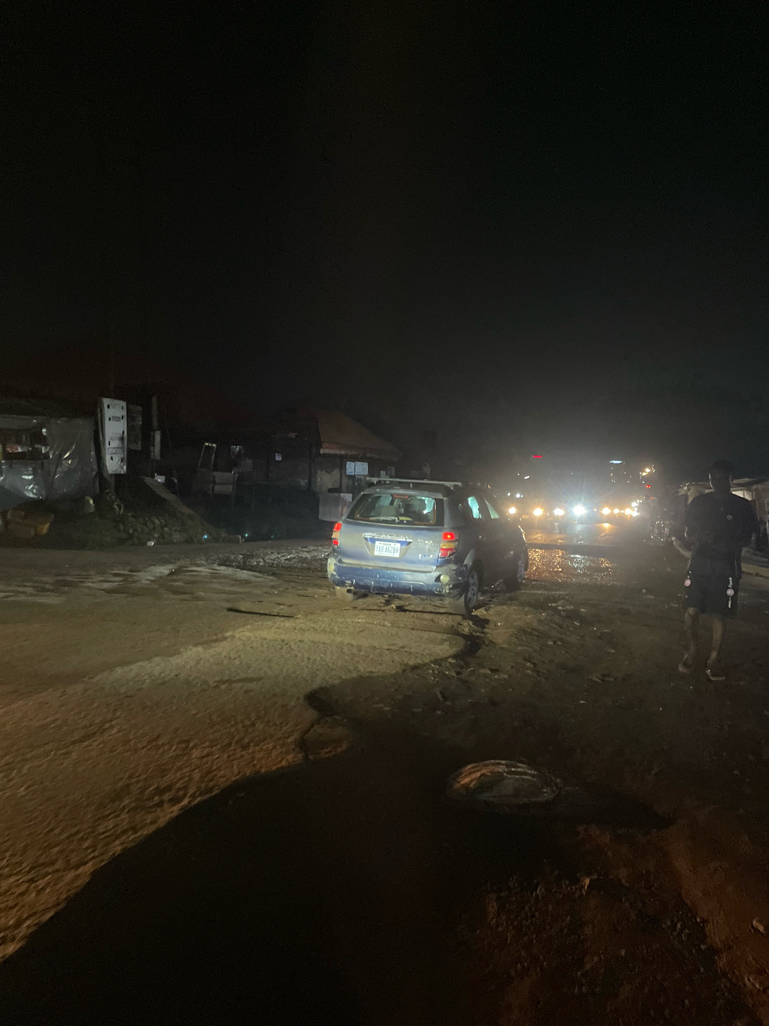 A blue SUV is parked on a dirt road at night in Abuja, Nigeria. The vehicle's license plate reads "YAB 462BH". The road is unpaved and appears muddy with puddles. To the left of the SUV are structures that resemble makeshift shops covered with tarps. In the background, there are more buildings and vehicles, with headlights from oncoming traffic illuminating the scene. A person is walking away from the camera on the right side of the road, wearing shorts and a t-shirt. The overall environment suggests a less developed or partially constructed area within the city.
