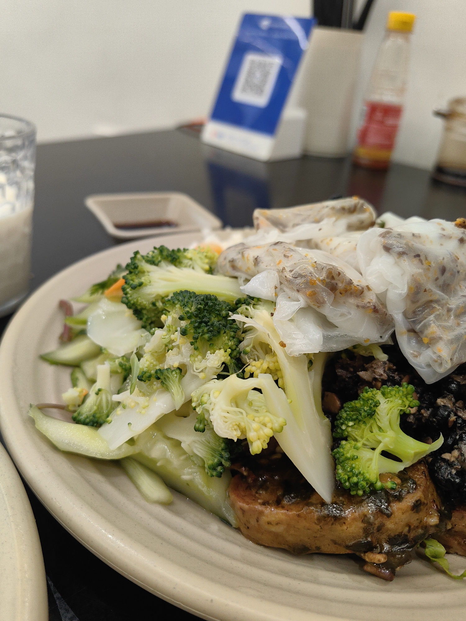 A plate of food, possibly spring rolls and vegetables, is in focus. The dish contains what appear to be Vietnamese spring rolls, accompanied by broccoli, other green vegetables, and dark, possibly spiced, ingredients. The plate is on a dark surface, likely a table. In the background, there is a clear glass with a liquid inside, a small container with a dark liquid (possibly soy sauce), a stand with a QR code on it, and a bottle with a yellow cap and red liquid inside. The scene is indoors, presumably a restaurant or dining area in Hong Gai Ward, Vietnam. There is no visible human interaction in the image.
