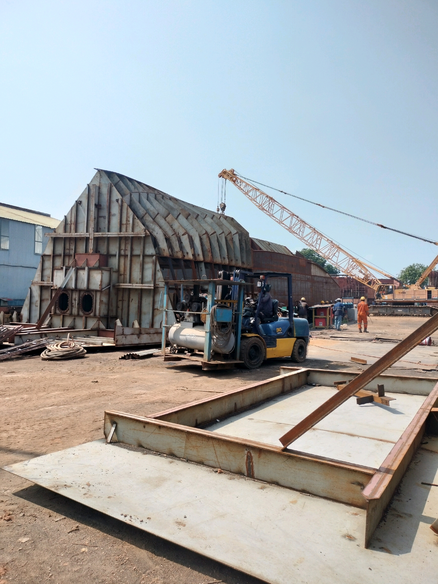 This image depicts an outdoor industrial construction site in Batam, Indonesia. The foreground features large, flat steel plates with a shallow, rectangular metal frame. In the mid-ground, a blue and yellow forklift is operated by a person wearing a dark uniform and a helmet. Adjacent to the forklift is a large, unfinished metal structure with a complex internal framing system and two circular openings on its side. A light blue compressor unit is positioned near the forklift. Further in the background, a yellow crawler crane is visible, along with another, larger crane with a yellow boom. Several workers in hard hats and orange jumpsuits are present, some standing and observing, while others appear to be engaged in work. The ground is predominantly dirt and gravel. The sky is clear and bright blue. The overall scene suggests ongoing shipbuilding or heavy industrial fabrication work.
