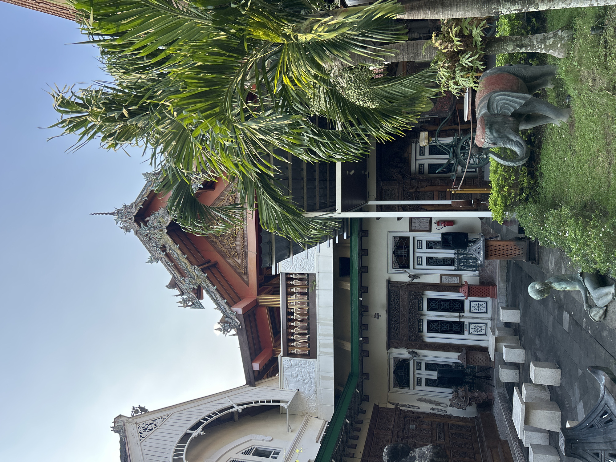 A medium shot captured from a slightly low angle shows the exterior of a building complex with traditional Indonesian architecture, located in Yogyakarta, Indonesia. The building features ornate carvings on its wooden doors and decorative elements on its roofs. A balcony with small statues of figures lines the upper level. In the foreground, a paved courtyard area is set with stone tables and benches. To the right, a green lawn is adorned with a stone elephant statue, and a statue of a seated child is visible on the left. Lush green foliage, including palm trees, surrounds the area. The sky is clear and blue.
