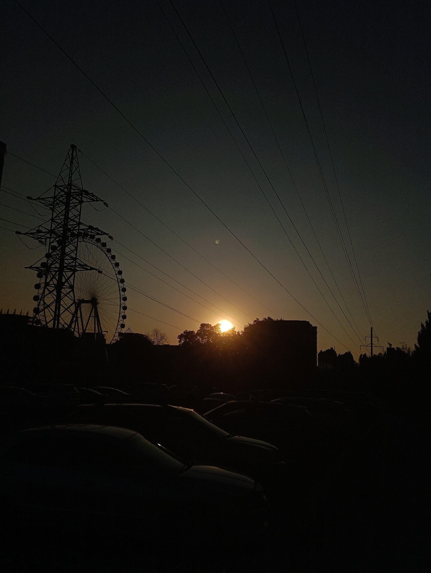 The image depicts a silhouetted cityscape at dusk. In the foreground, rows of parked cars are visible, their shapes mostly obscured by shadow. A large power line tower stands prominently to the left, with a Ferris wheel partially visible behind it, also in silhouette. The sun is setting behind trees and buildings in the mid-ground, casting a warm glow across the sky, with visible power lines crisscrossing the upper portion of the frame. The overall atmosphere is that of an urban setting as day transitions into night.
