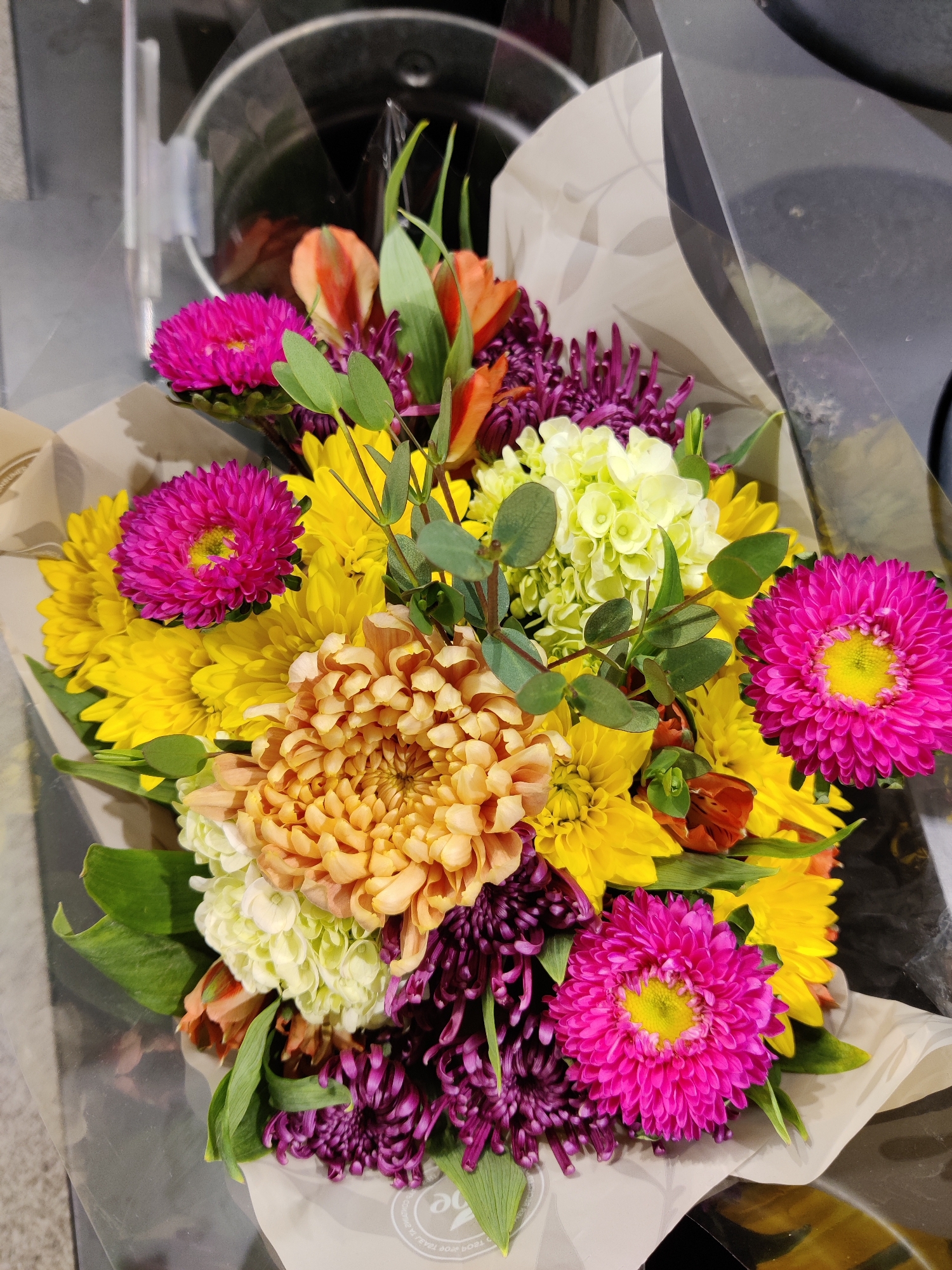 A bouquet of flowers is displayed in a clear plastic wrapper. The bouquet contains a variety of flowers including bright pink asters, yellow chrysanthemums, orange alstroemeria, light green hydrangeas, and purple spider mums. The flowers are arranged tightly together, with some green foliage interspersed. The bouquet is resting on a light brown paper wrap, and a portion of a white circular logo is visible on the wrap. The background is blurred, but appears to be a retail environment with metallic surfaces. The scene is likely located in Mechanicsburg, United States of America.