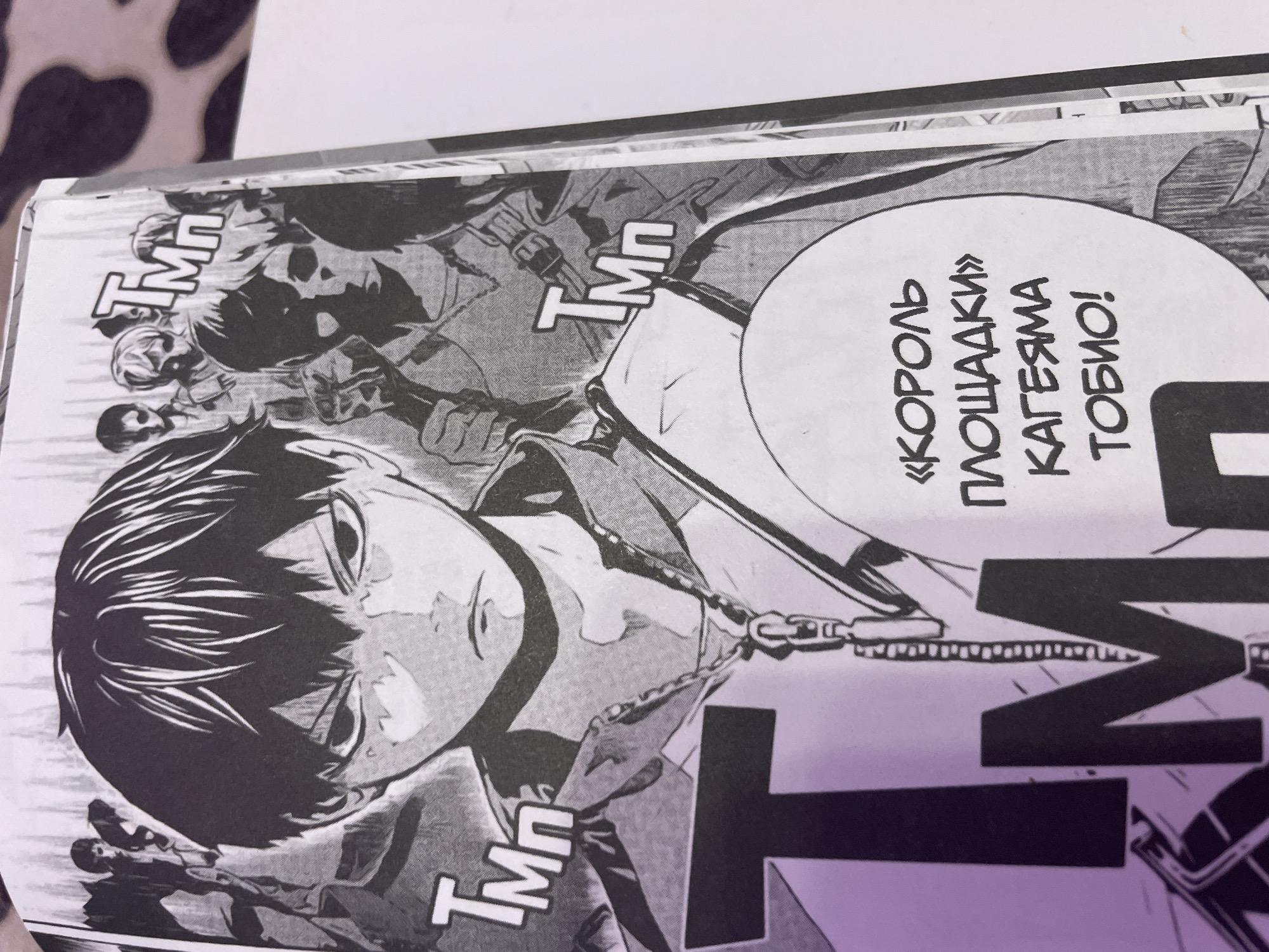 The image is a close-up of a manga panel featuring an anime character with spiky black hair and an intense expression, looking directly at the viewer. The character is wearing a jacket with a zipper and a chain. In the background, other anime characters are visible, some with their arms raised. There are also large, stylized text elements that appear to be sound effects or labels within the manga. A speech bubble contains text in a Cyrillic alphabet. The image appears to be a page from a manga book. There is no discernible information or depiction related to the city of Irinovka, Russia.
