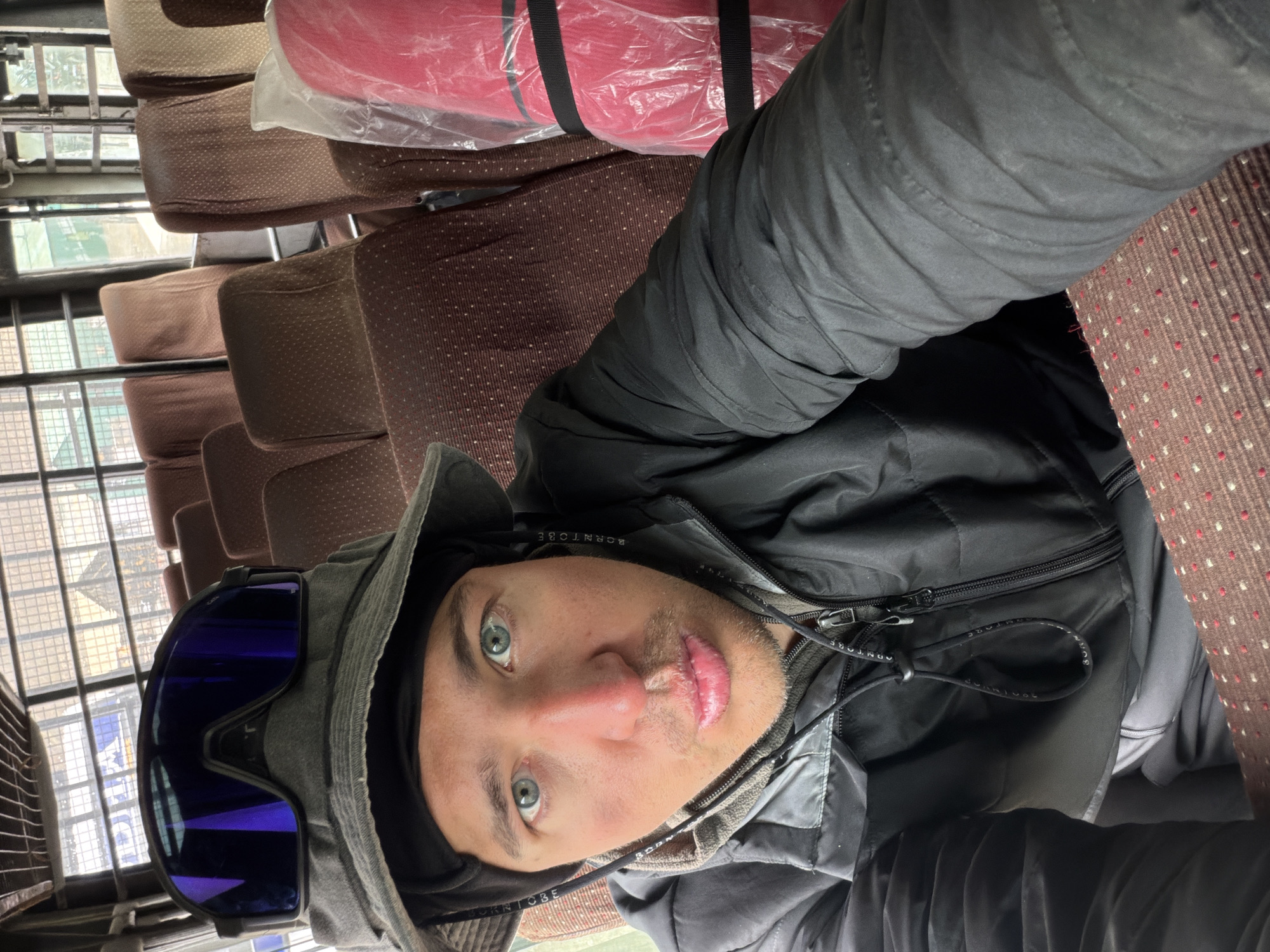A close-up, selfie-style photograph shows a person wearing a grey bucket hat and black ski goggles, with the goggles' lenses reflecting a purple hue. The person has blue eyes and is looking directly at the camera with their lips slightly parted. They are wearing a black jacket over a grey hoodie and are seated in a vehicle with rows of brown, patterned seats. To the right of the person, a red object wrapped in clear plastic and secured with black straps is visible. The background shows a window with a metal grate and hints of greenery outside. The overall lighting suggests an indoor setting, likely during the daytime. The image appears to have been taken on a smartphone.