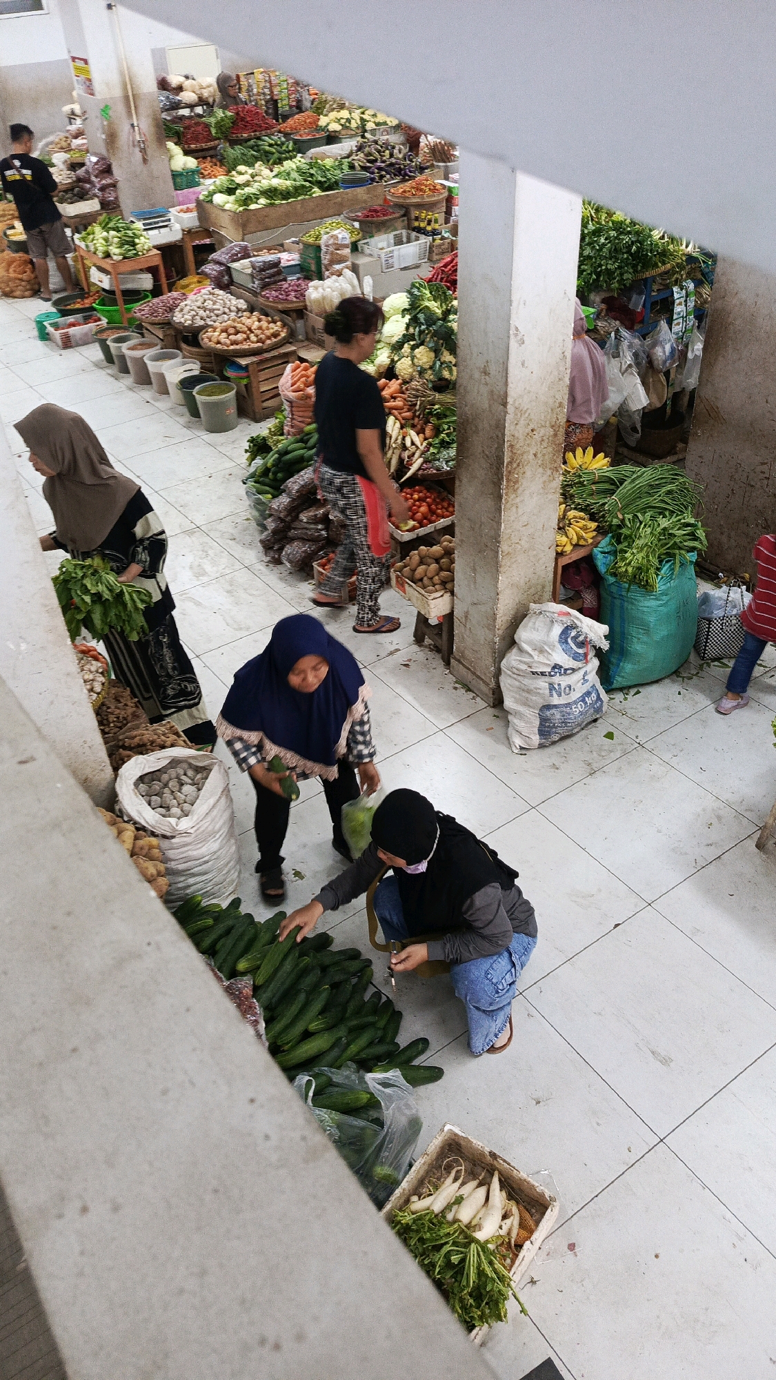 The image depicts an outdoor market scene in Nologaten, Indonesia, characterized by a tiled floor and an abundance of fresh produce. Several individuals are engaged in activities related to the market. In the foreground, two women are observed interacting with cucumbers. One woman, wearing a blue hijab and a plaid top, is holding a cucumber and appears to be selecting vegetables. The other woman, in a black hijab and blue jeans, is crouched down and also handling cucumbers. Behind them, a woman in a black hijab and patterned dress carries a bunch of green leafy vegetables. Further back, a person in a black t-shirt and shorts is visible. The market stalls are filled with various fruits and vegetables, including what appear to be carrots, cauliflower, tomatoes, onions, potatoes, bananas, and other leafy greens. Bags of produce, some made of woven material and others of plastic, are scattered around the scene. The overall impression is one of a bustling local market in Indonesia, with people actively participating in the buying and selling of fresh goods.