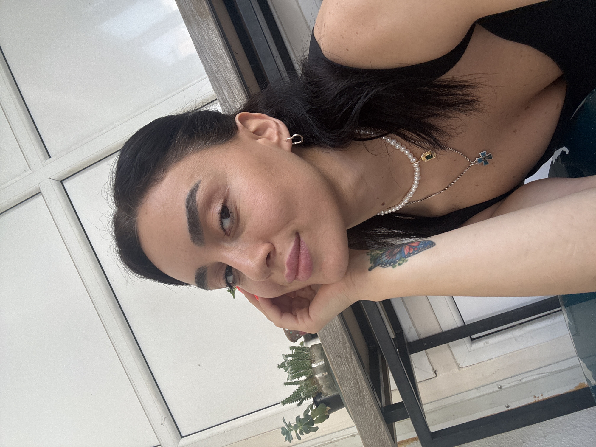 A young woman with dark hair is taking a selfie outdoors, leaning her head on her hand. She is wearing a black top, a pearl necklace with a pendant, and another necklace with a blue cross pendant. There is a tattoo of a butterfly on her left forearm. In the background, there are several potted plants on a shelf and a window with white frames. The scene appears to be on a balcony or patio of a building in Tbilisi, Georgia, suggested by the architectural style of the window frames and the outdoor setting.