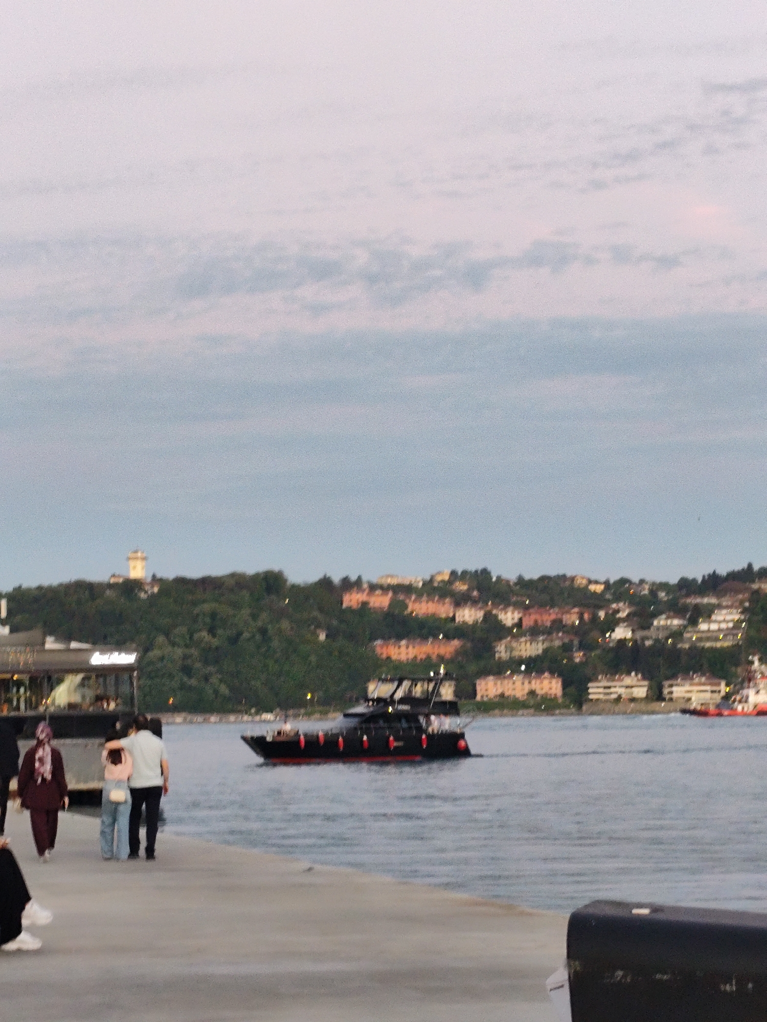 The image depicts a waterfront scene in Sarıyer, Turkey, likely taken at dusk given the soft light and pastel sky. A couple walks arm-in-arm along a concrete pier or walkway, and a woman in a maroon headscarf and clothing walks in front of them. To the left, there is a section of a building. A black boat with red trim is on the water, moving away from the pier. Across the water, a tree-covered hill is covered in buildings and houses and a white tower stands at its peak. The water is calm, with ripples on the surface. Another boat is seen in the distance, to the right. A partially visible black object is in the lower right corner.