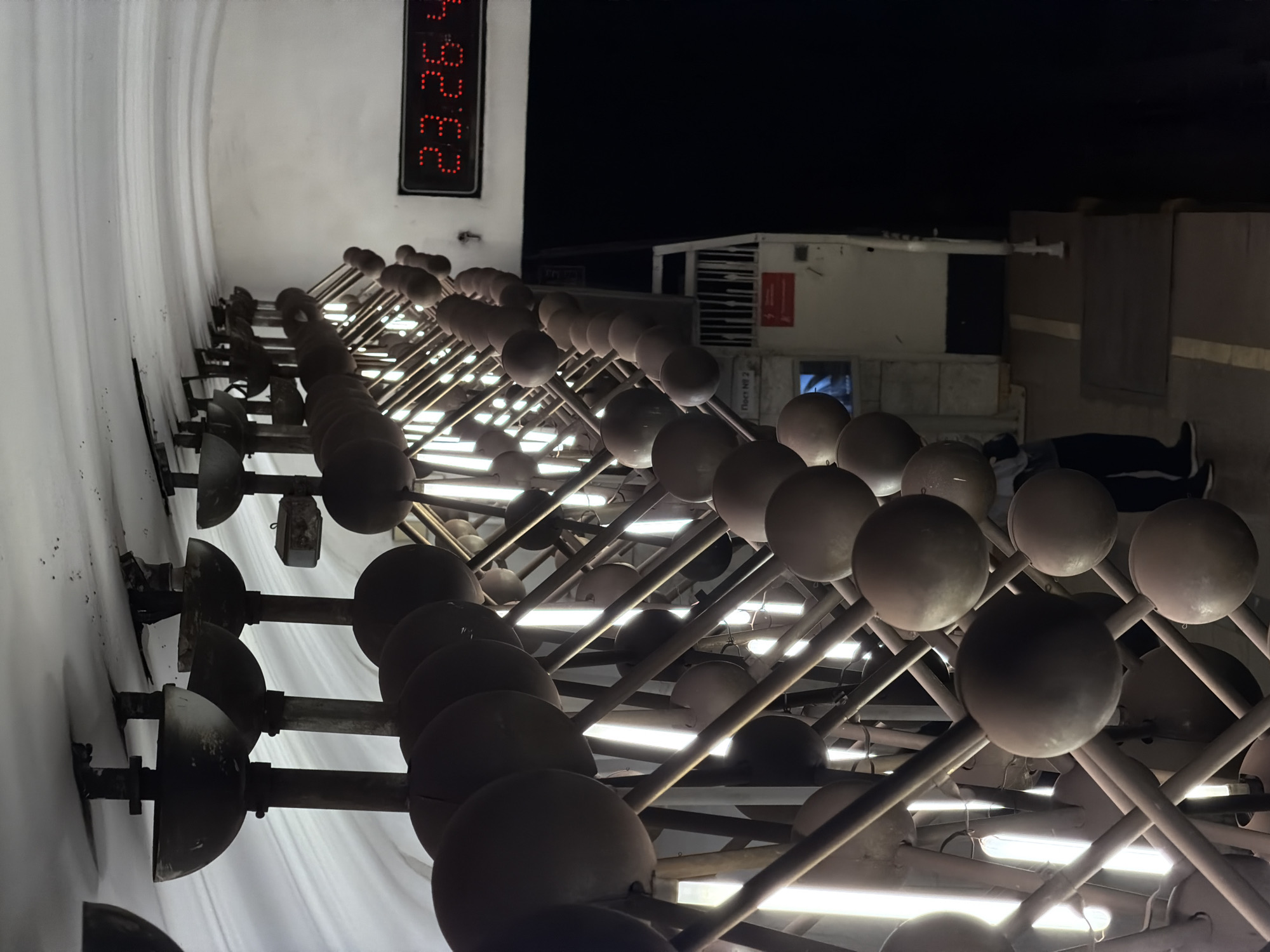 The image shows a nighttime view of the "Atom" pavilion at VDNKh, a large exhibition park in Moscow, Russia. The photo is taken from a low angle, looking up at the pavilion's facade, which features a decorative design of spheres attached to rods, arranged in a grid pattern. Linear lights are integrated into the grid, illuminating the structure. A digital clock showing "23:26" is visible on the building's facade. In the background, other buildings within VDNKh can be seen. A person is lying on the ground in the periphery of the frame.